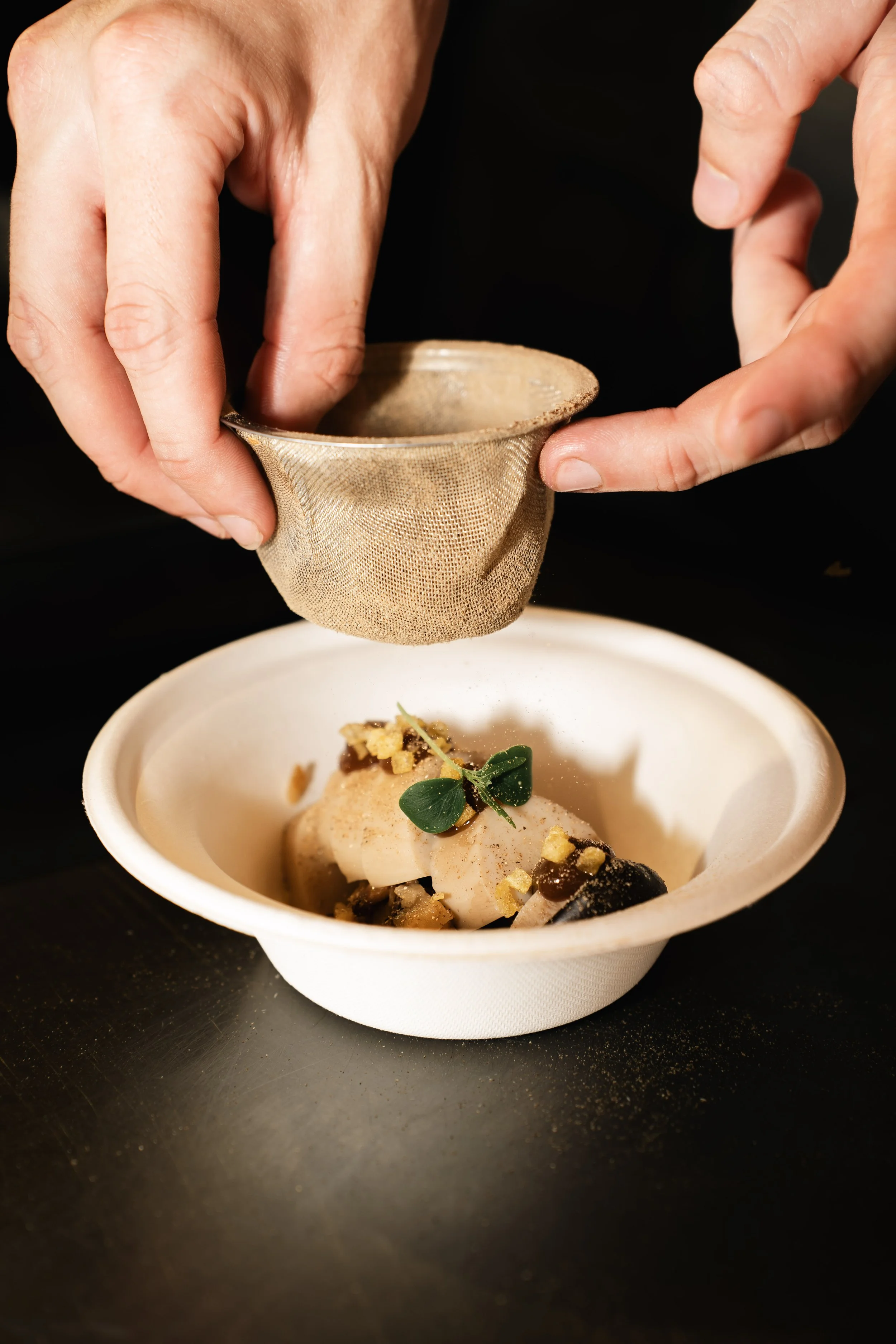 Klein bord met een gerecht, bedekt met een klein Kruidentje, en er ligt een stuk vis met saus en toppings, terwijl een persoon een goudbruine zeef of vergiet boven het gerecht houdt.