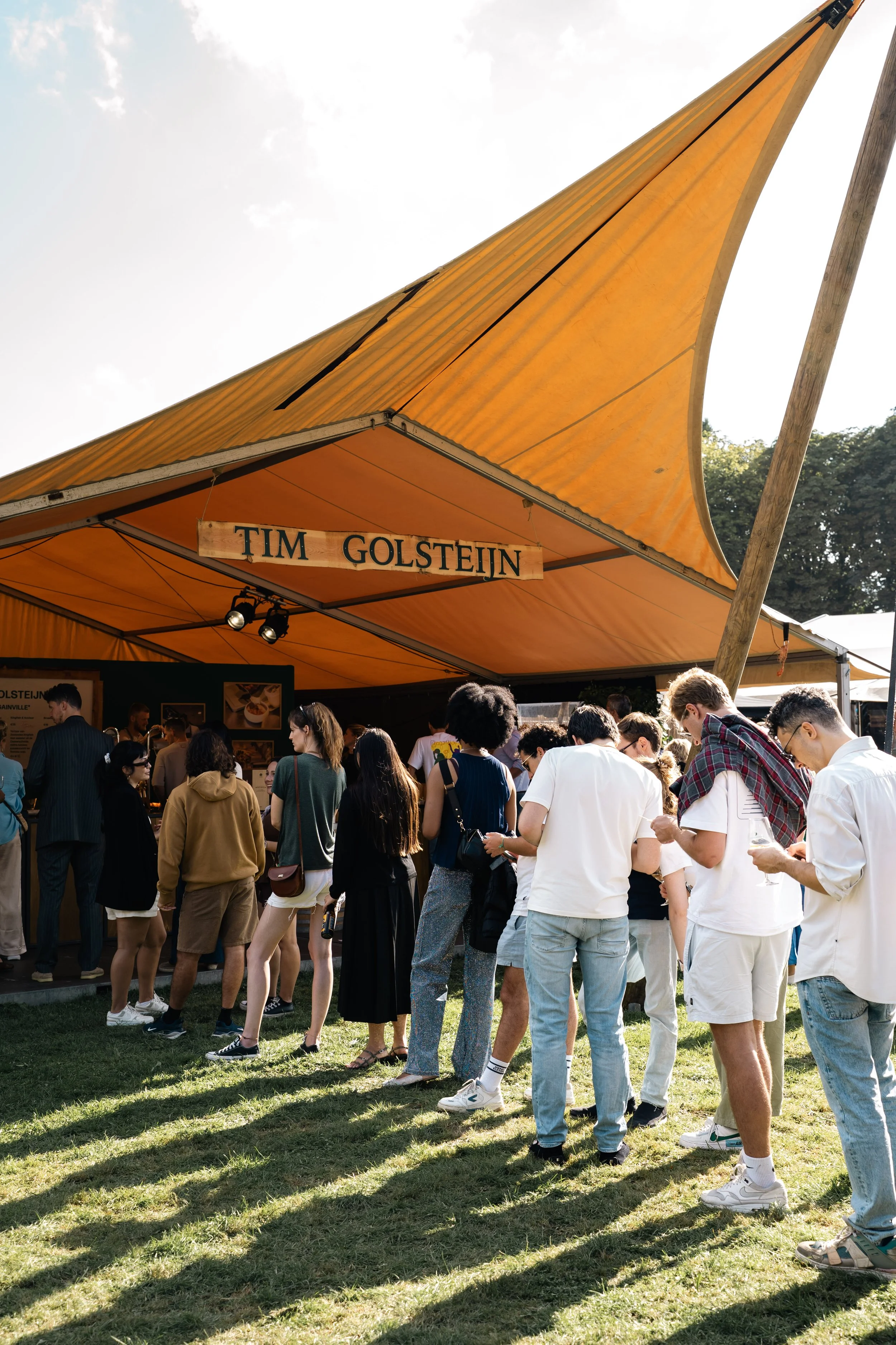 Een groep mensen wacht in de rij onder een oranje tent, met een bord met de tekst 'TIM GOLSTEIN'.