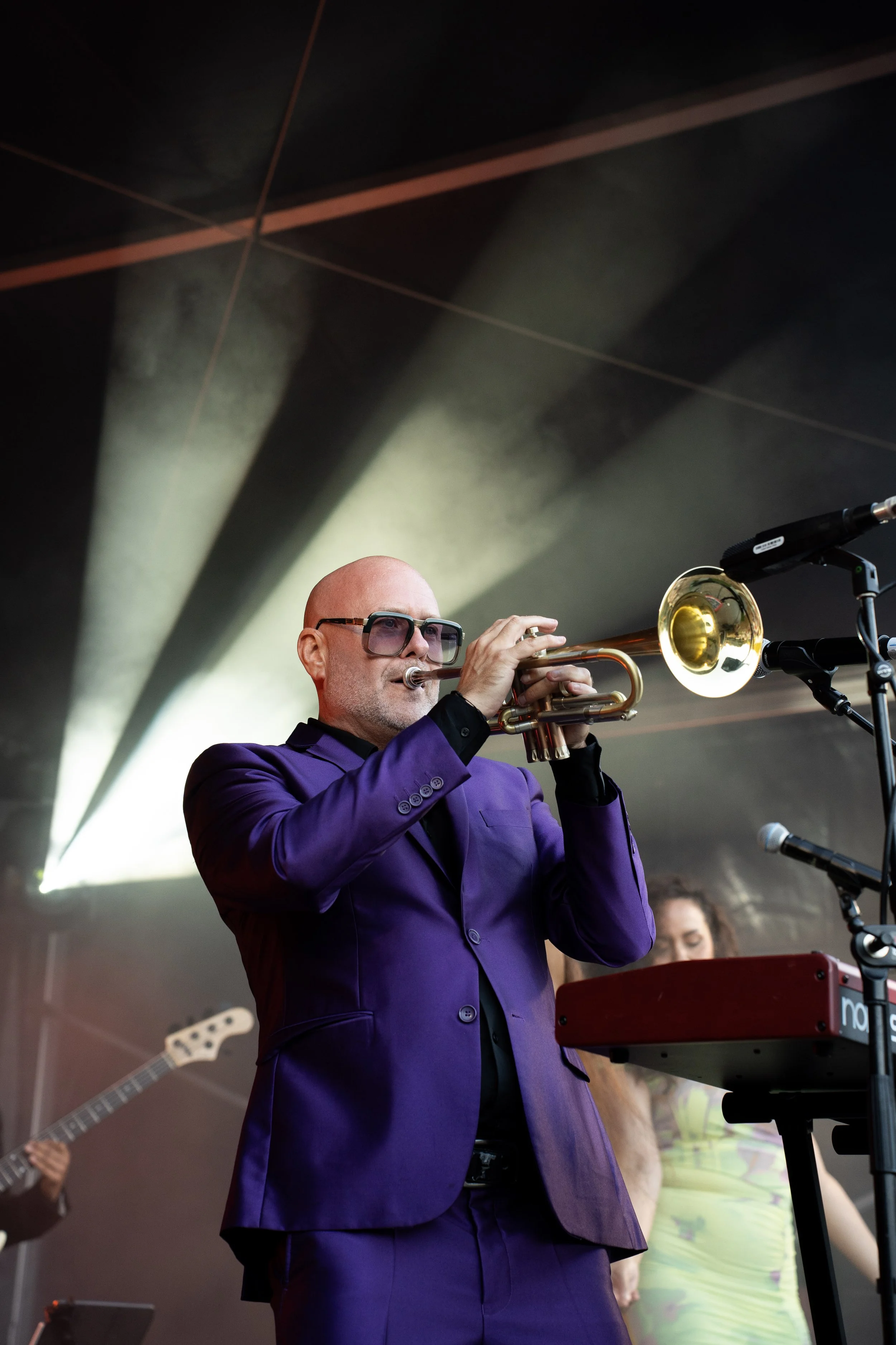 Mannelijke muzikant in een paarse pak speelt trompet tijdens een optreden, met andere muzikanten op de achtergrond.