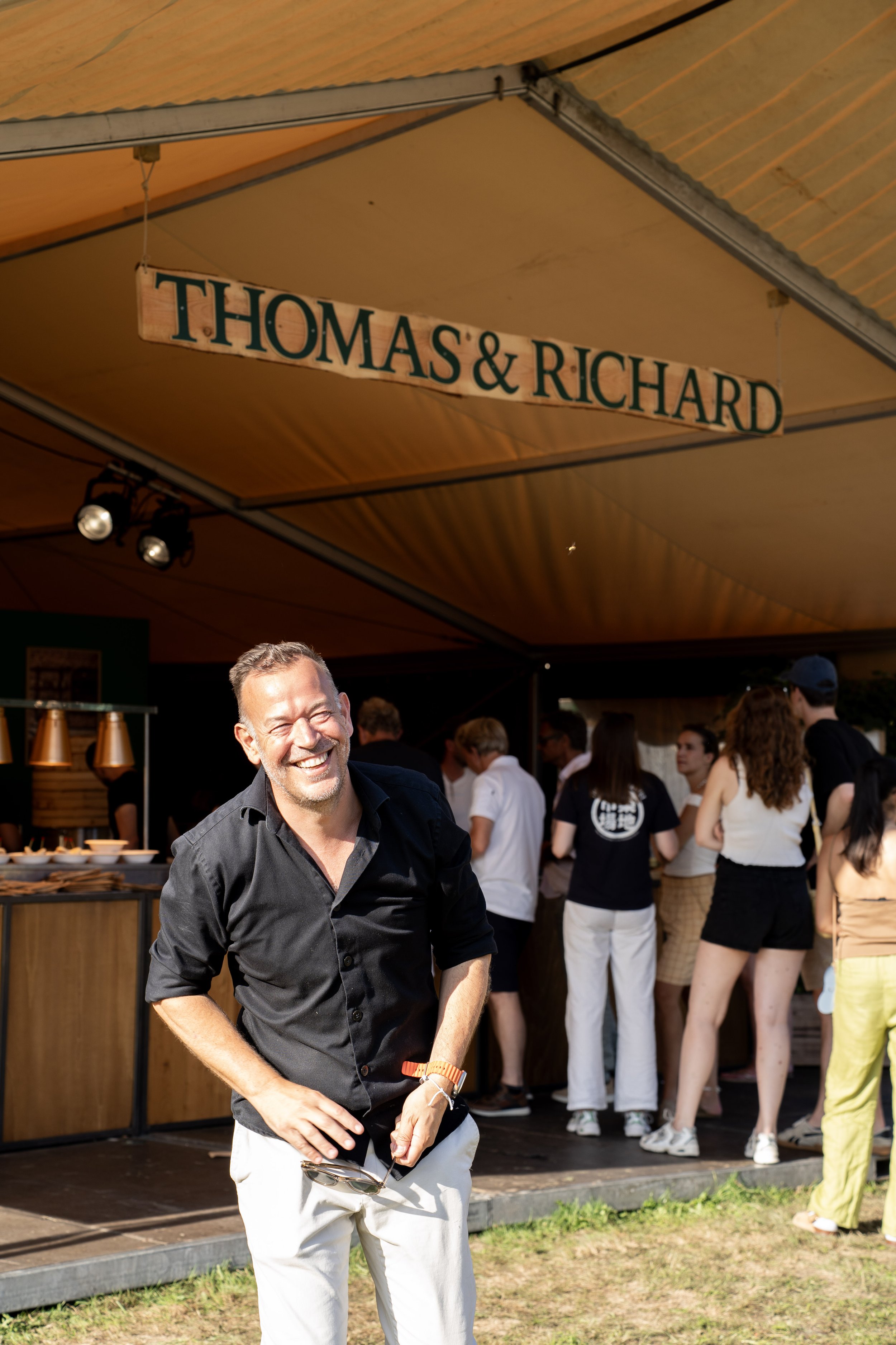 Een lachende man met een zwart overhemd en witte broek staat voor een groep mensen onder een oranje tent met een bord dat 'Thomas & Richard' zegt. Mensen staan in een rij bij een buffet, waarschijnlijk op een buitenfeest of evenement.