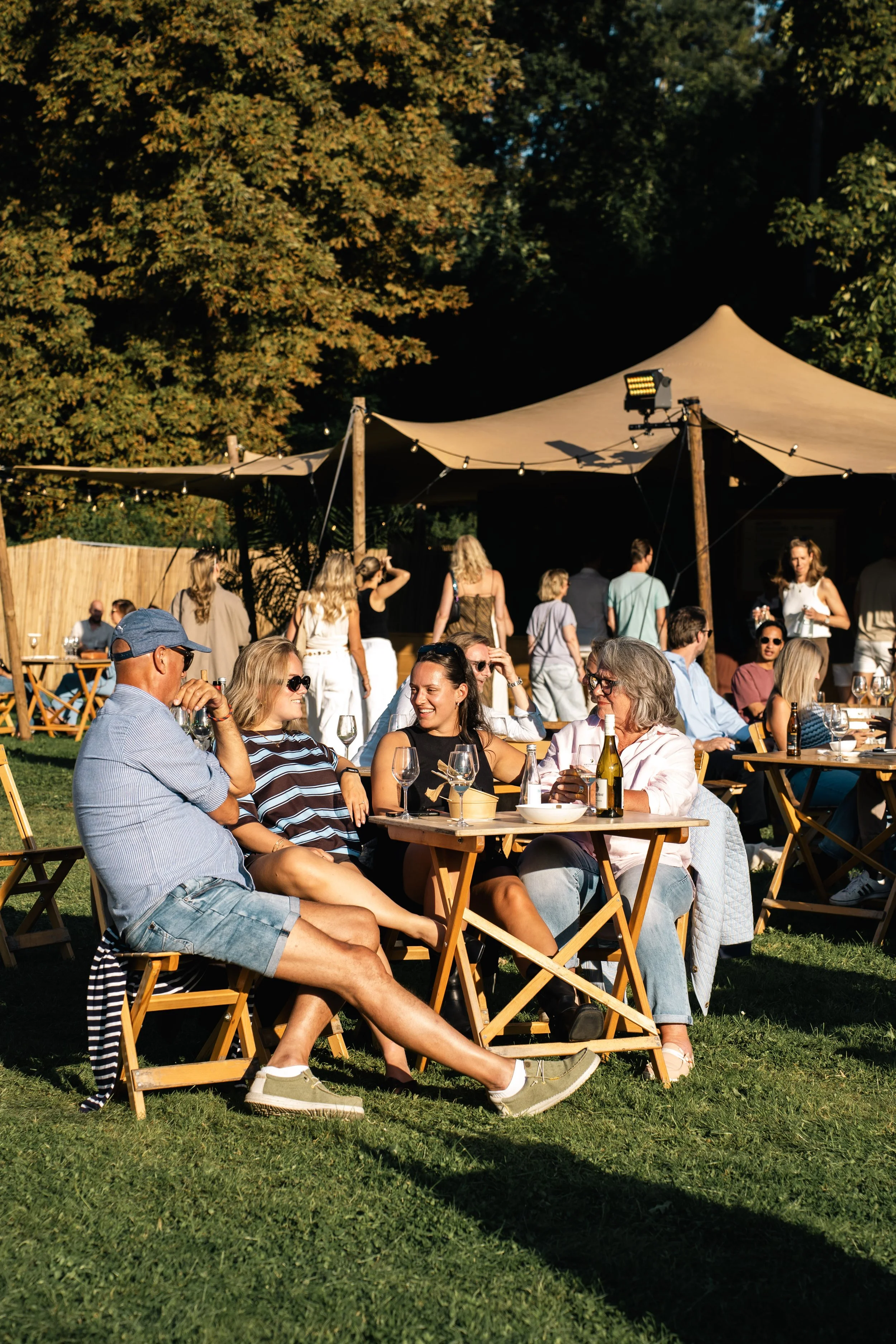 Een groep mensen die buiten op houten stoelen zit en plezier heeft tijdens een zomerse dag, met een tent boven hen en grote groene bomen op de achtergrond.