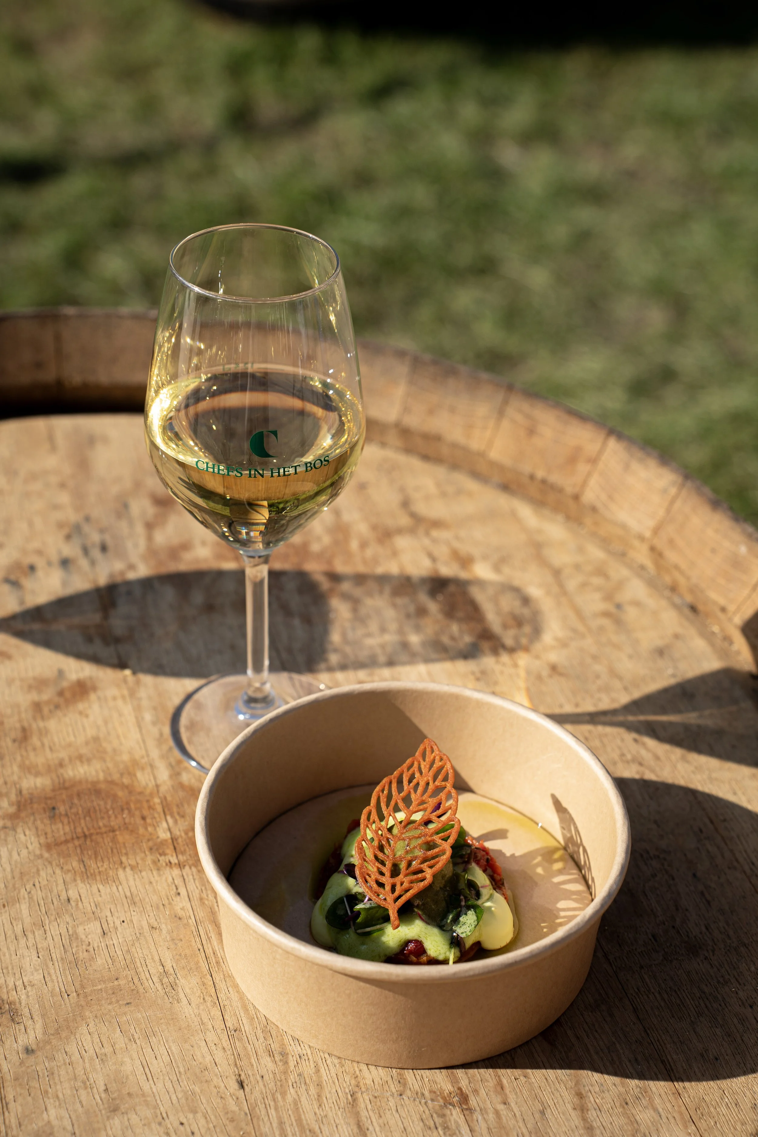 Glas witte wijn op een houten tafel met een bordje met voorgerecht in de voorgrond, in een groene buitenomgeving.