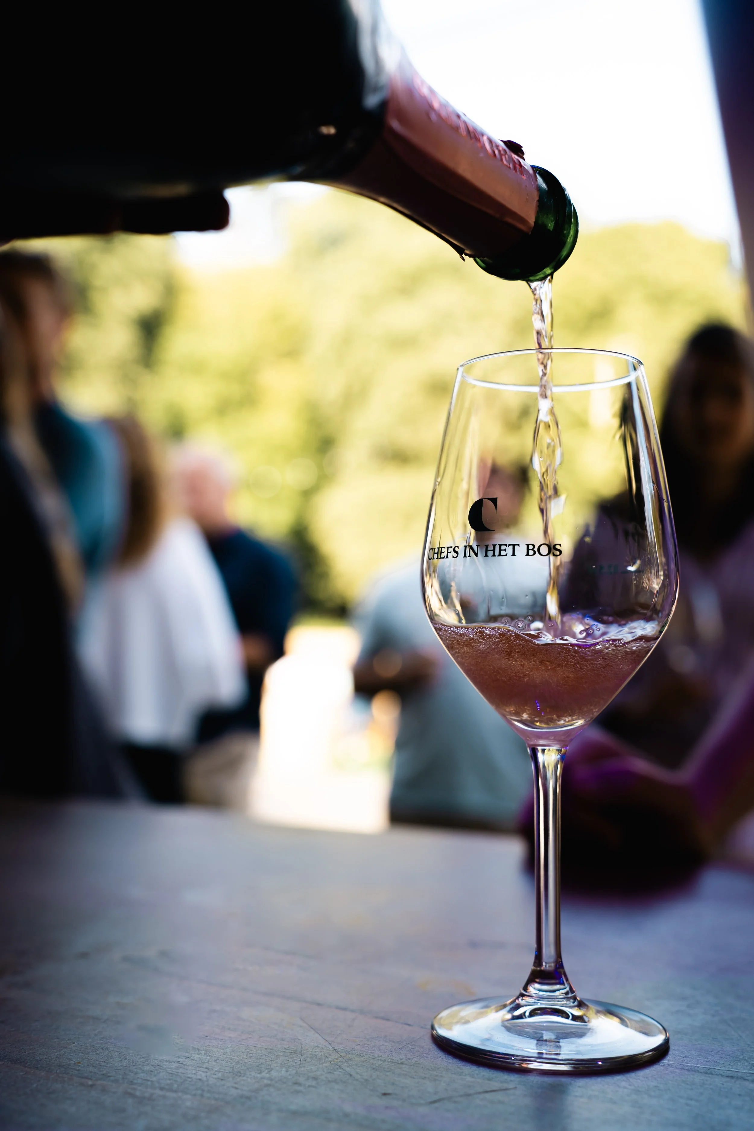 Een wijnglas met pinkrosé wijn, met de tekst 'chefs in het bos' op het glas, op een houten tafel, met mensen op de achtergrond die uitkijken naar een buitengebeurtenis.