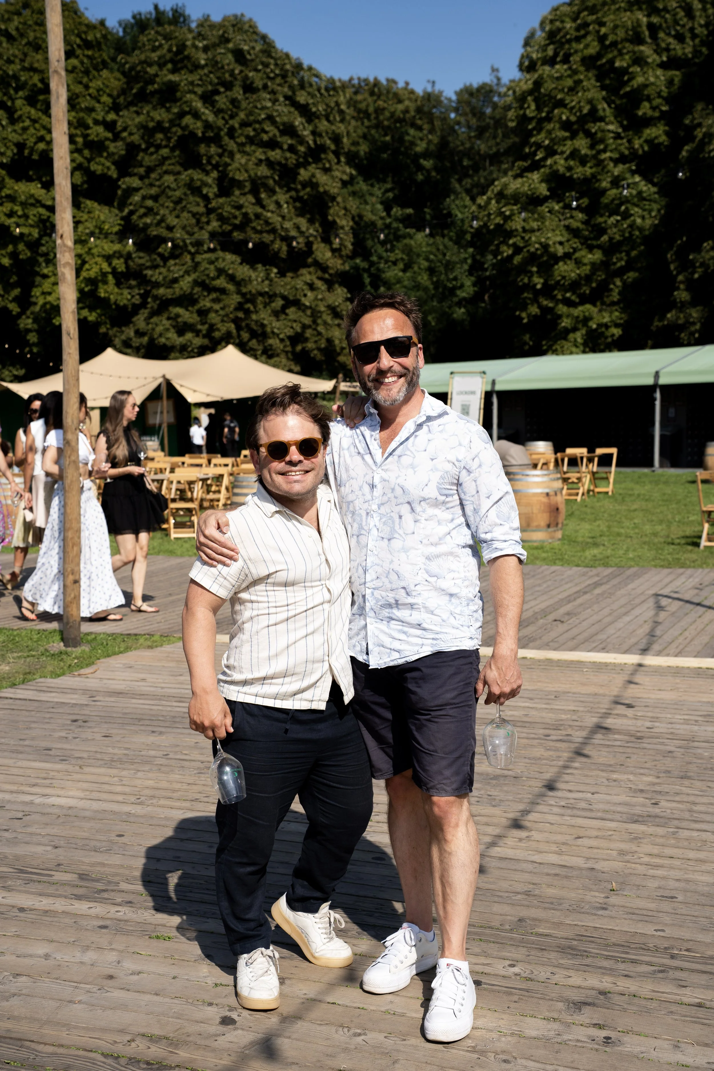 Twee mannen in zomerkleding glimlachen en staan samen op een houten terras tijdens een zomerevenement, met mensen en tenten op de achtergrond.