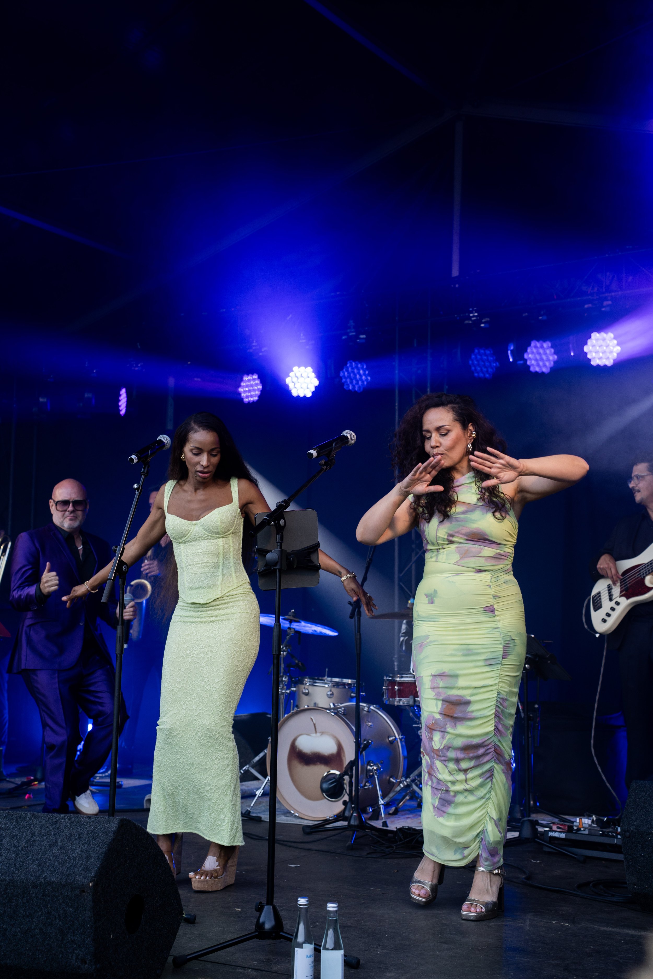 Twee vrouwen dansen en zingen op een podium tijdens een live muziekoptreden, met muzikanten en instrumenten op de achtergrond, onder blauwe en paarse lichtstralen.