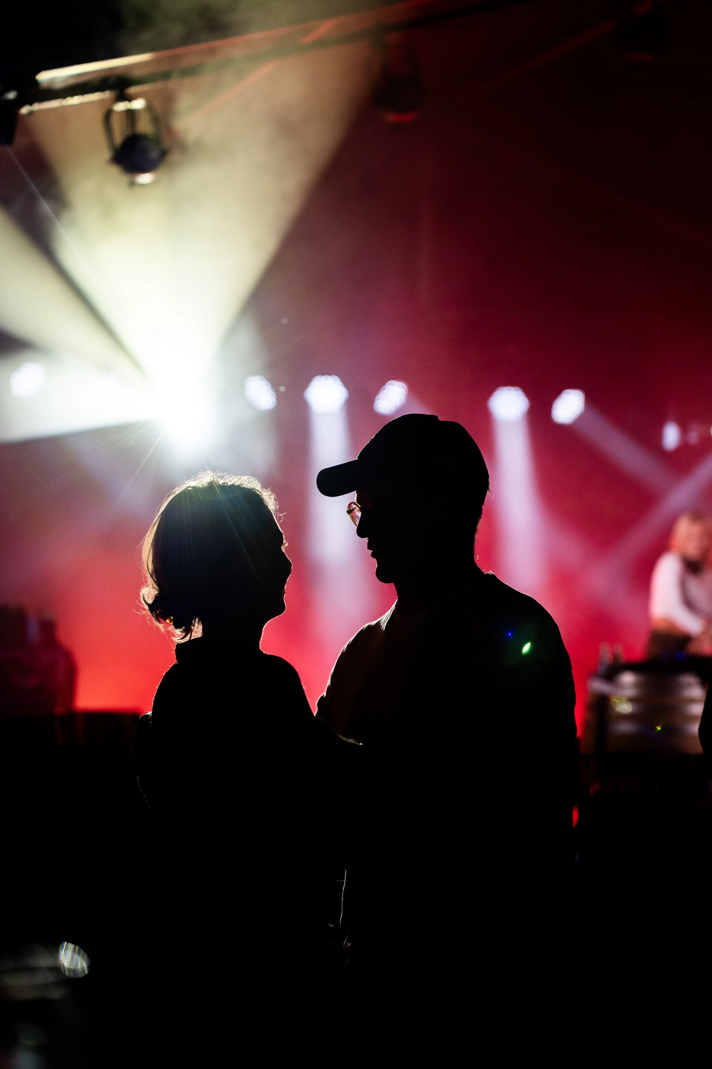 Silhouetten van een man en een vrouw die samen kijken in een donkere omgeving met rood en wit licht, mogelijk bij een muziek- of dansfeest.