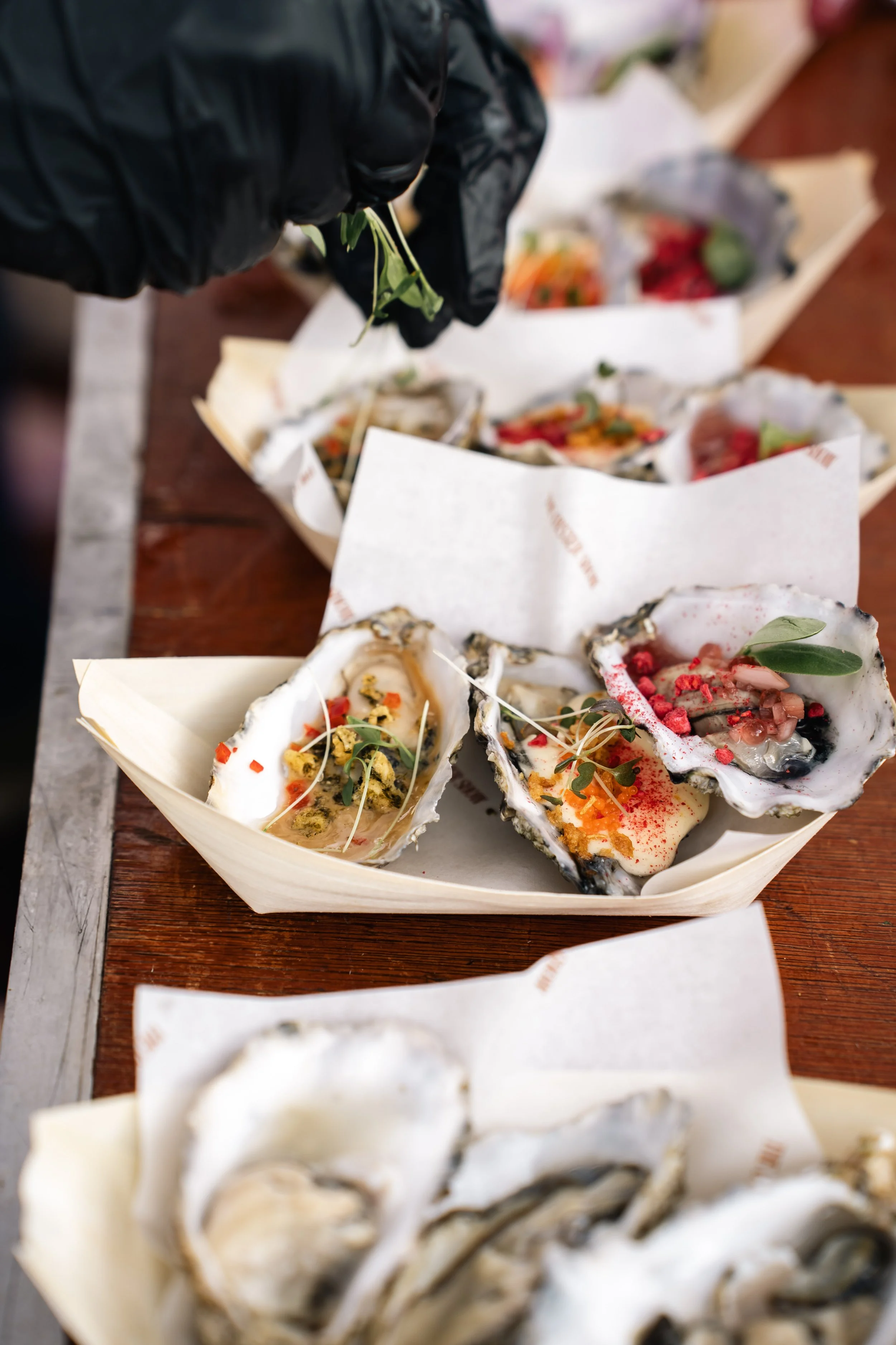 Close-up van verschillende oysters in bedrukte papieren schaaltjes op een houten tafel, versierd met groenten en kruiden.