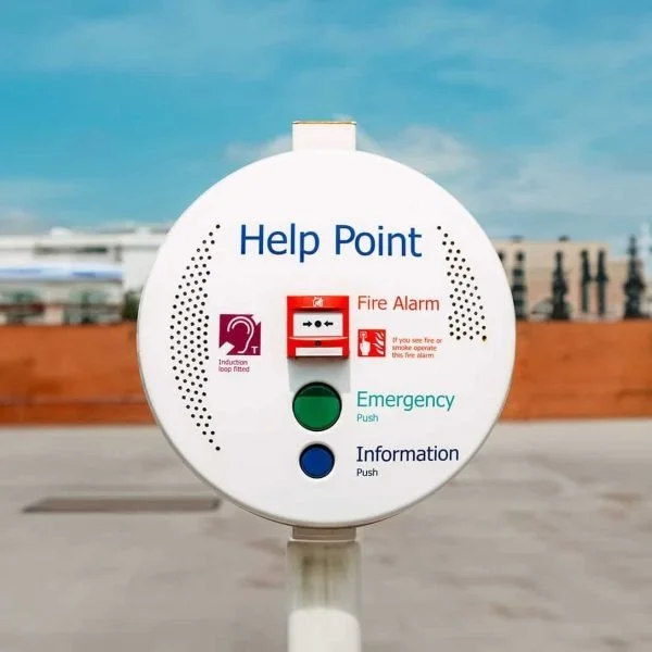 A large, circular help point station with a fire alarm button, an emergency button, and an information button, set against a blurred outdoor background.
