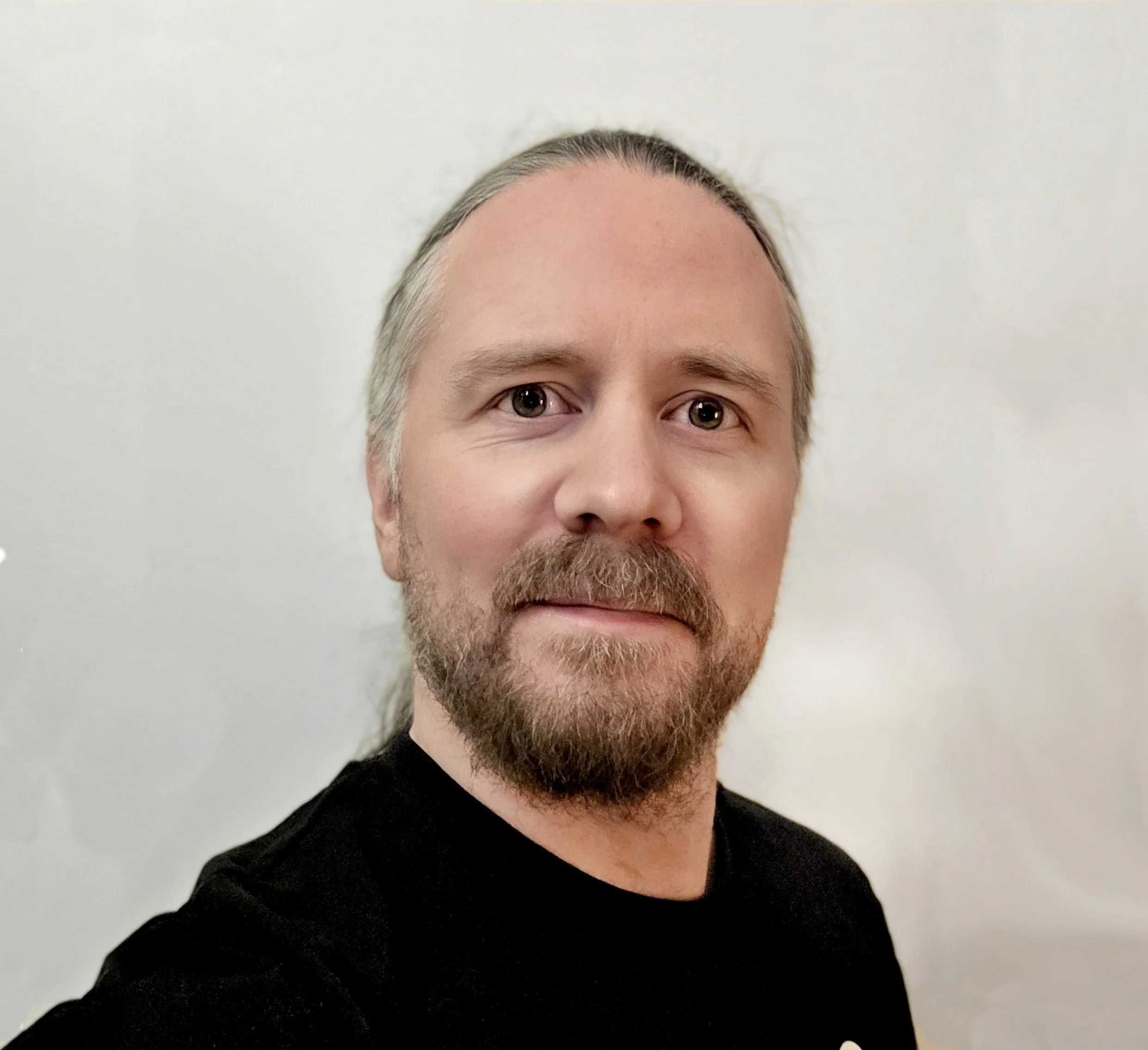 Portrett av en mann med langt brunt hår, skjegg og mustasje, iført en svart t-skjorte med en Alice Cooper-logo.