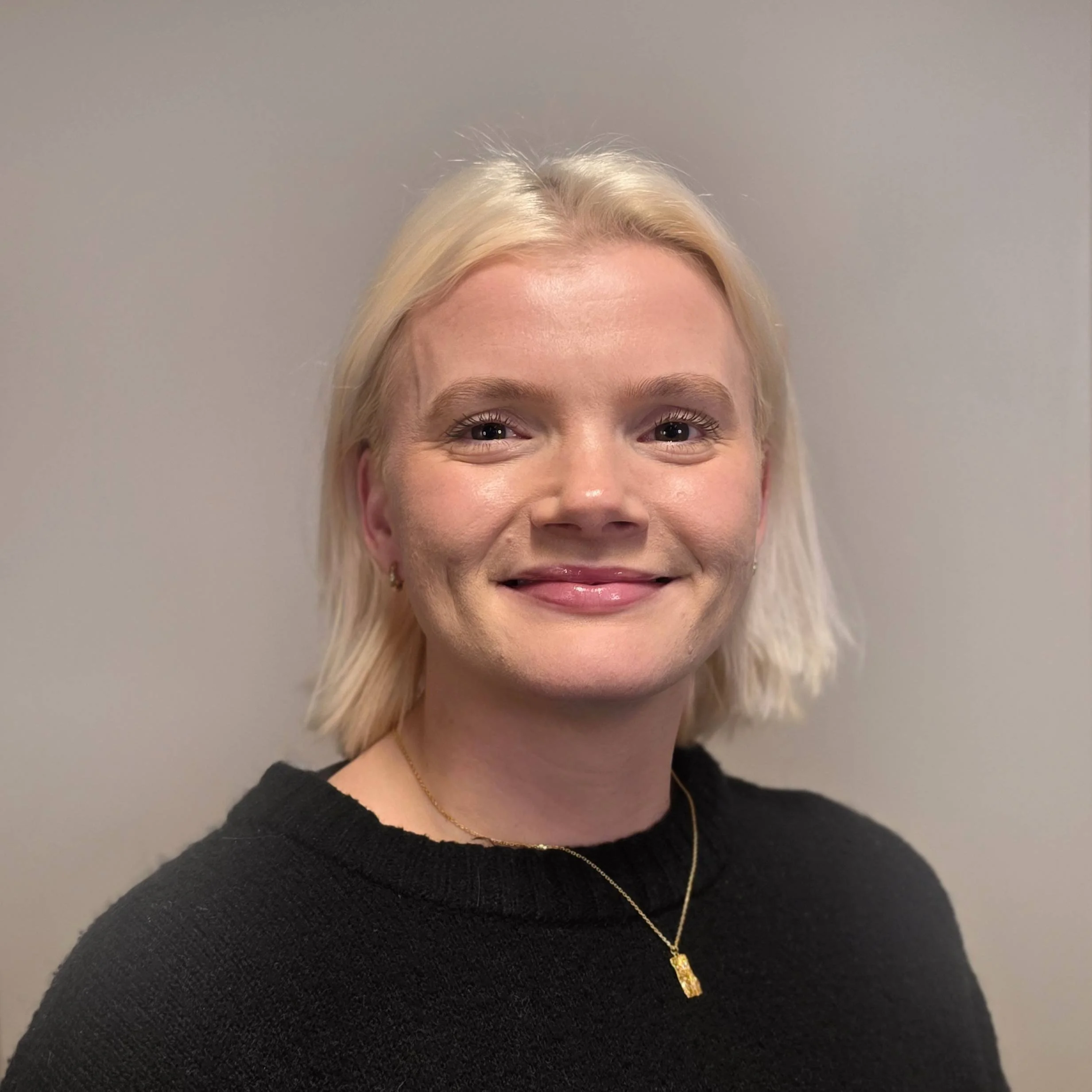 Kvinne med lys hud og blondt hår smilende og iført svart turtleneck, mot en lys bakgrunn.