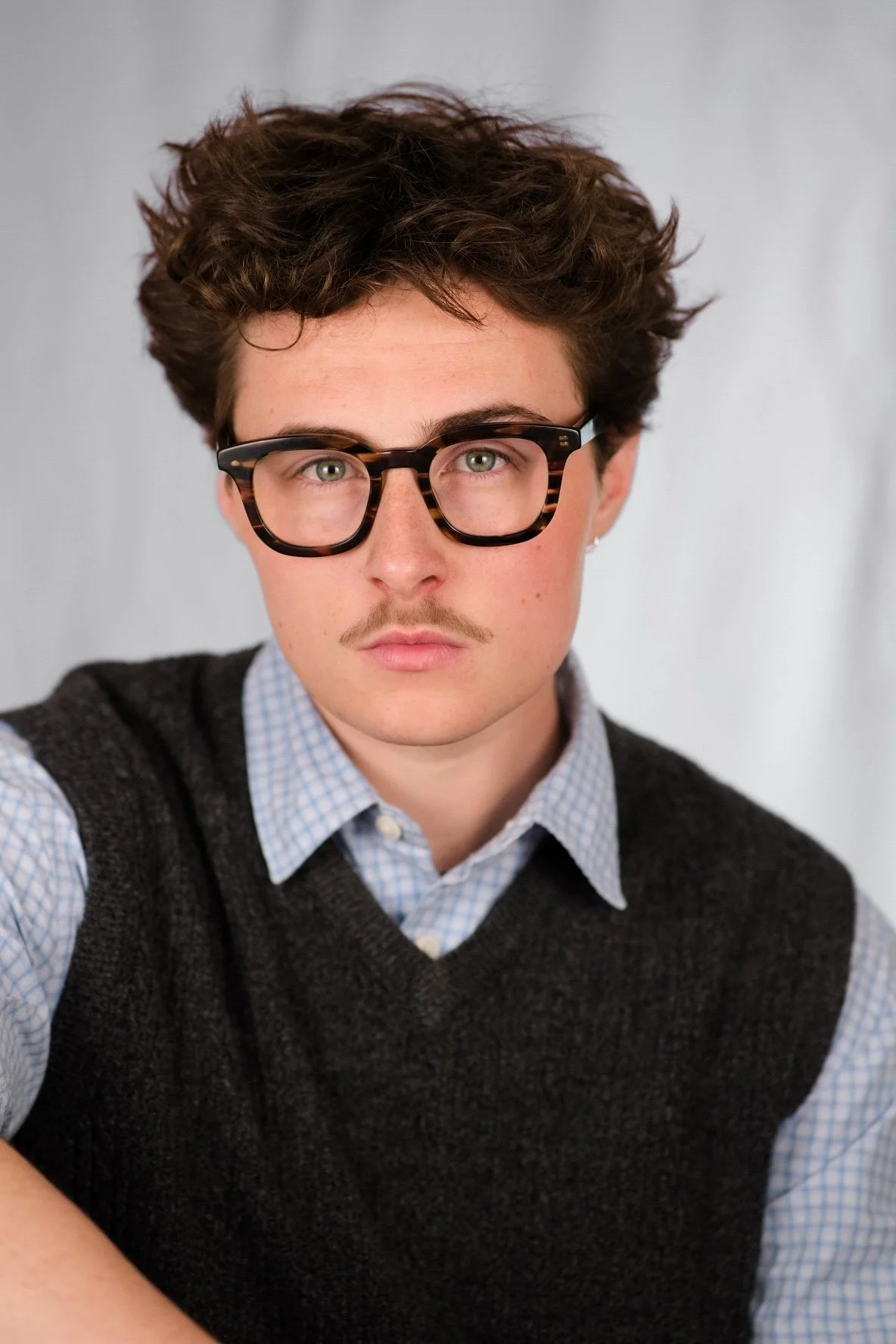A young man with curly brown hair, wearing glasses with tortoise shell frames, a light blue checkered shirt, and a dark vest, looking directly at the camera against a light gray background.