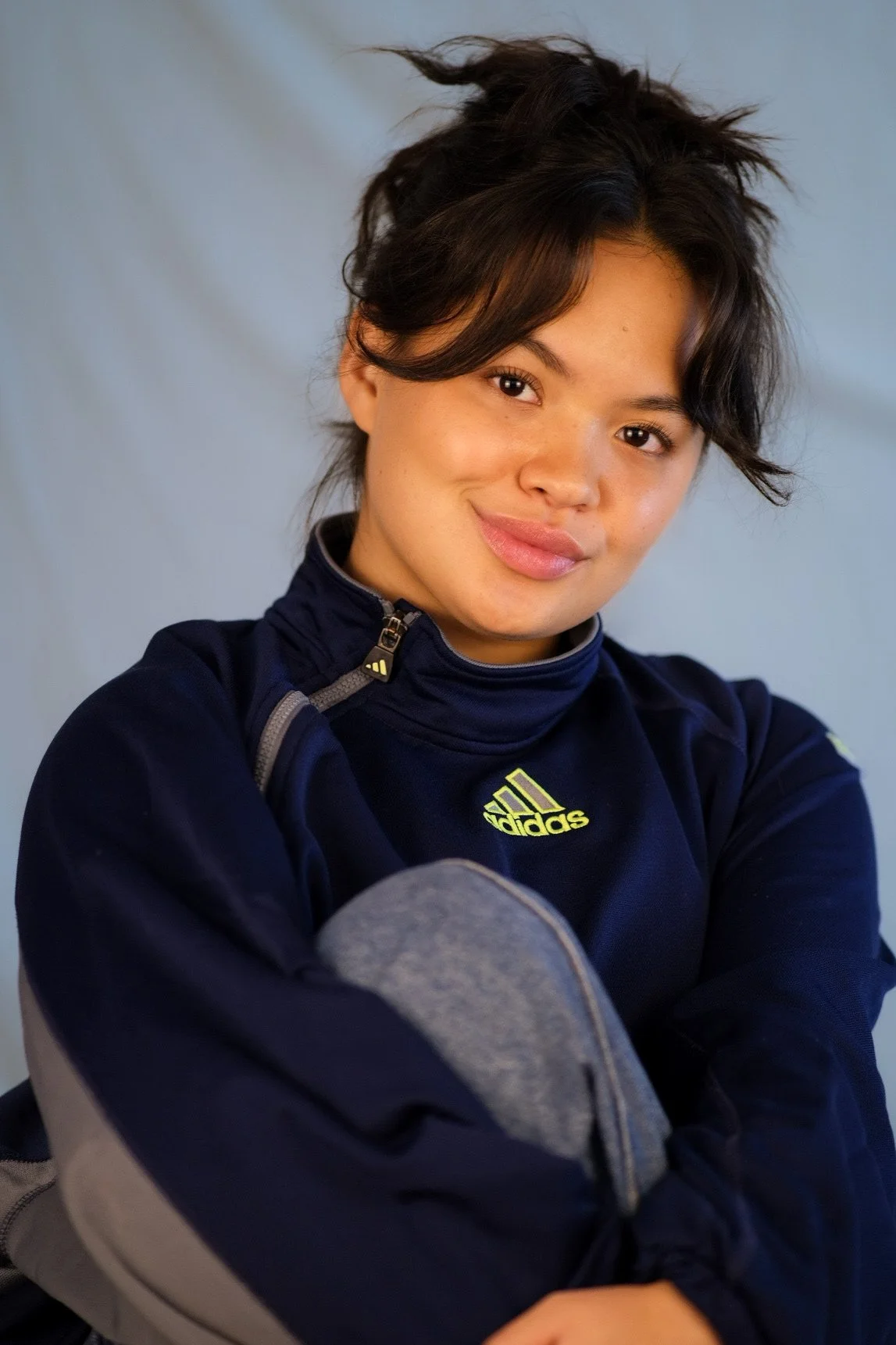 A young woman with short, messy black hair, smiling, wearing a navy blue sporty jacket with a yellow Adidas logo, crossing her arms, against a plain gray background.