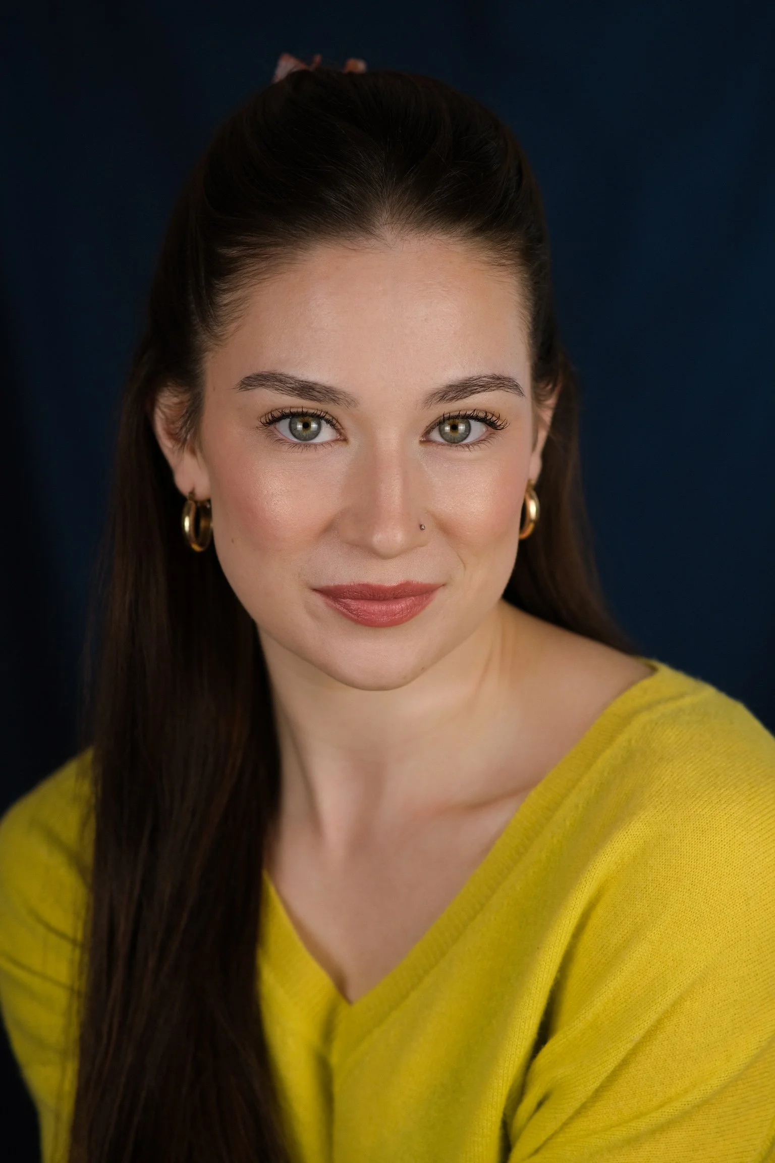 Portrait of a woman with greenish-gray eyes, wearing a yellow sweater and gold hoop earrings, with long dark brown hair, against a dark background.