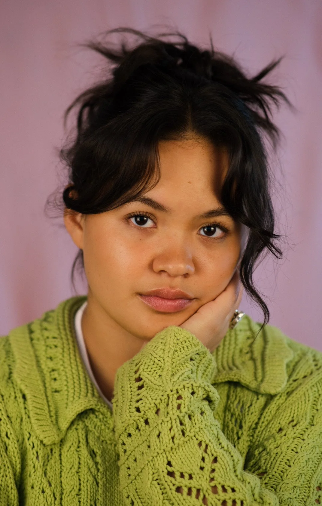 A young woman with dark wavy hair and tan skin, wearing a green knitted sweater, looking at the camera with her hand resting on her face, against a pink background.