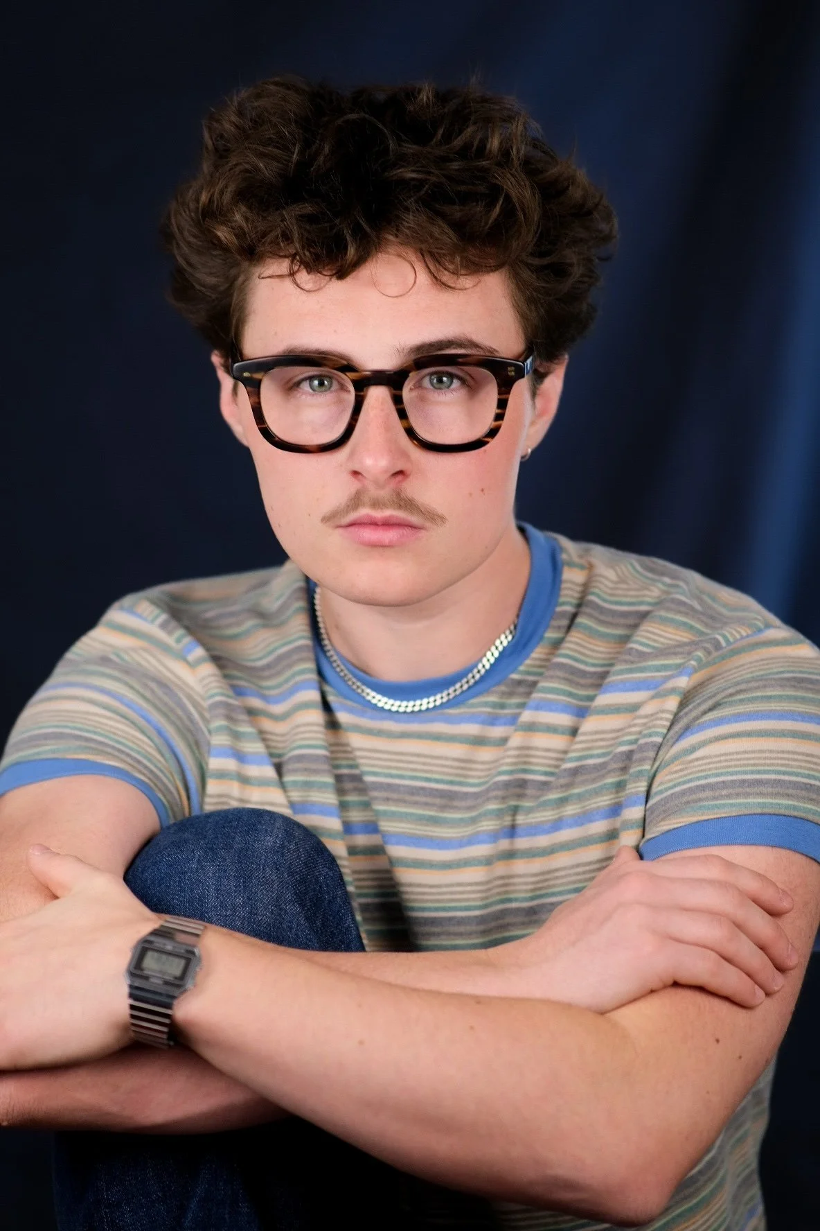 Portrait of a young man with curly brown hair, glasses, and a mustache, wearing a striped T-shirt, jewelry, and a watch, sitting with arms crossed against a dark background.