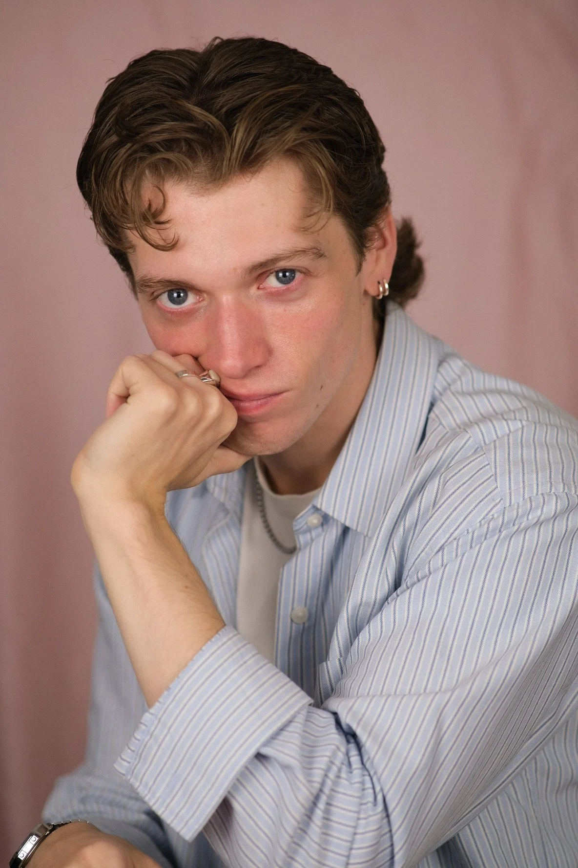 A young man with brown hair, blue eyes, and multiple earrings, wearing a light blue striped shirt, resting his chin on his hand and looking into the camera with a neutral expression, against a pink background.