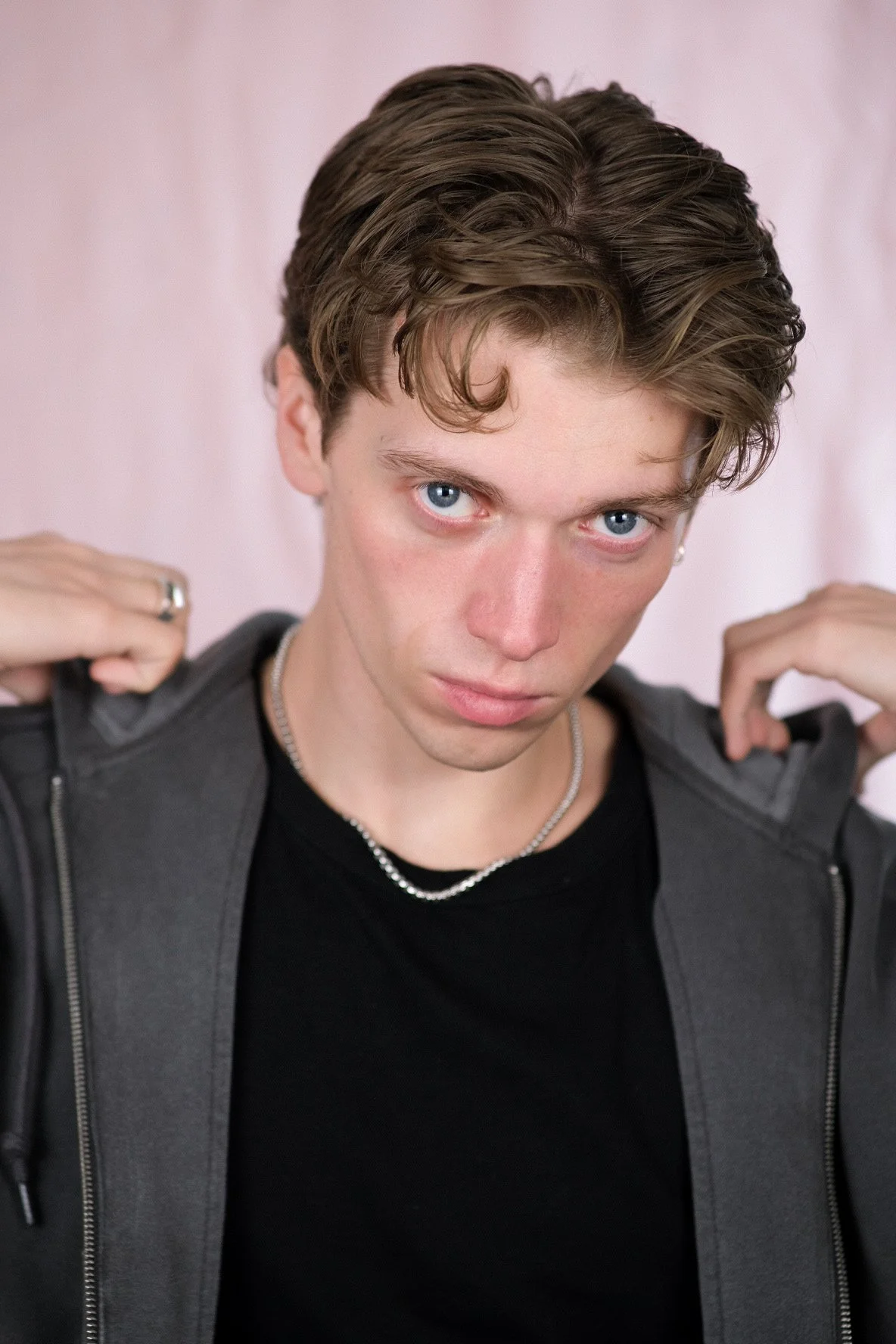 A young person with short, wavy brown hair and blue eyes looking directly at the camera, wearing a black shirt, a grey jacket, and silver jewelry, holding the hood of the jacket with both hands, standing against a light pink background.