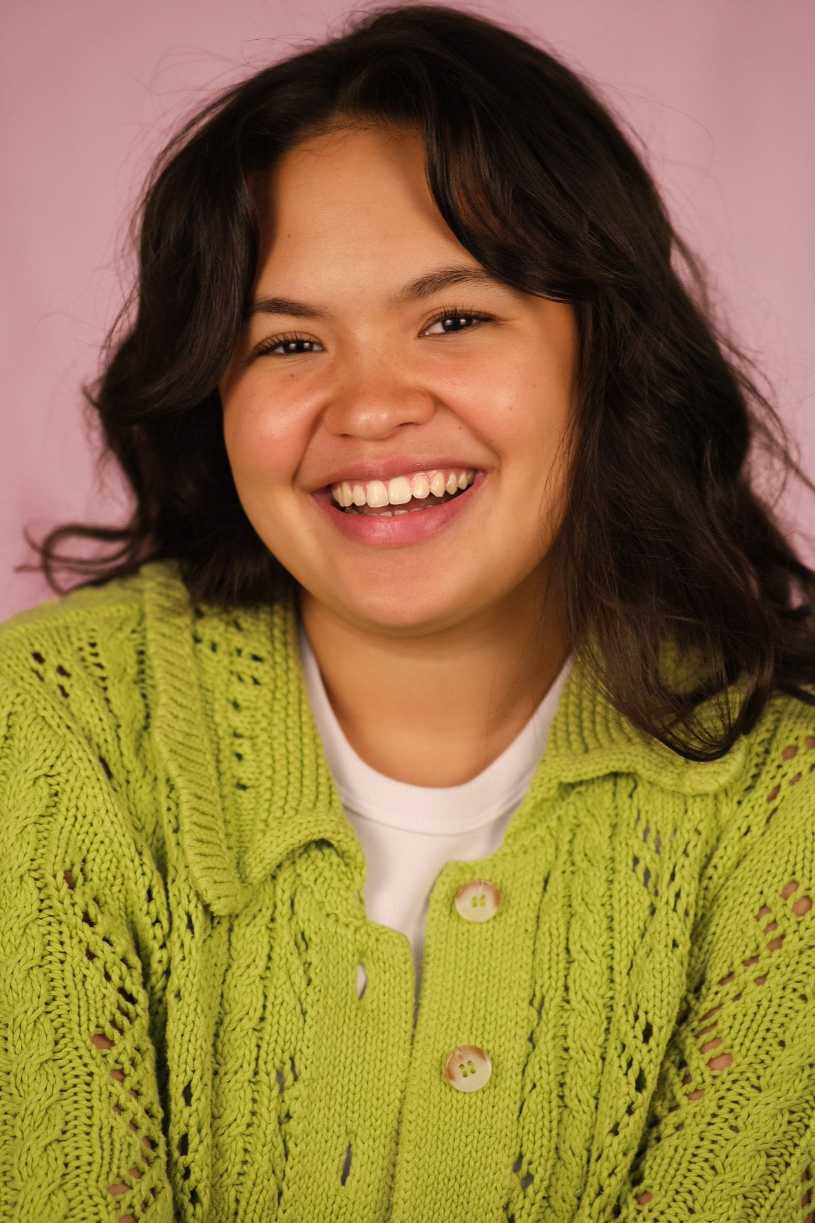 A young woman with dark curly hair smiling, wearing a bright green knit cardigan over a white shirt, against a pink background.