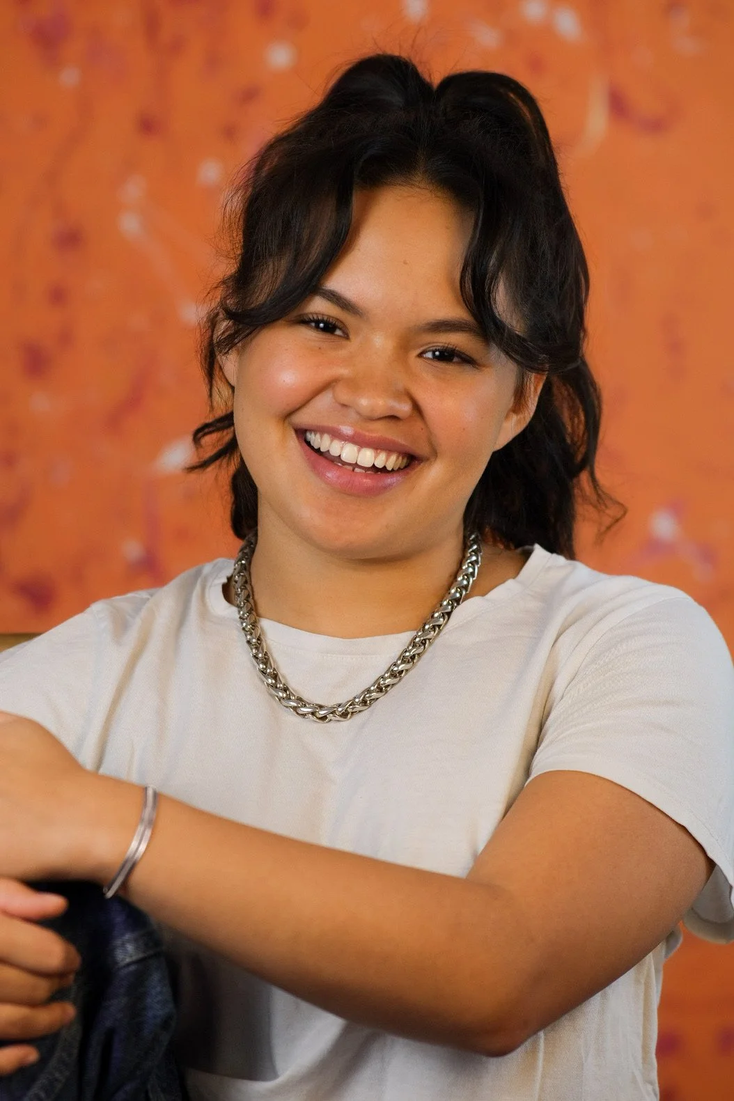 A young woman with shoulder-length dark hair, smiling, wearing a white t-shirt, a silver chain necklace, and a bracelet. The background is orange with patterns.