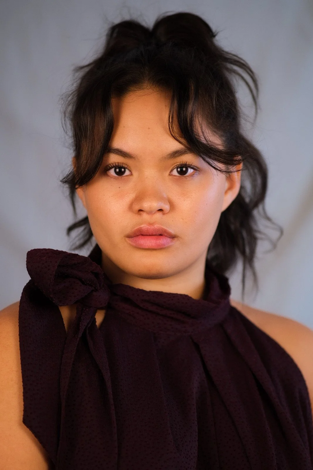 Close-up photo of a young woman with dark hair styled in loose waves, wearing a dark purple or black sleeveless top with a tied bow on her shoulder, set against a plain background.
