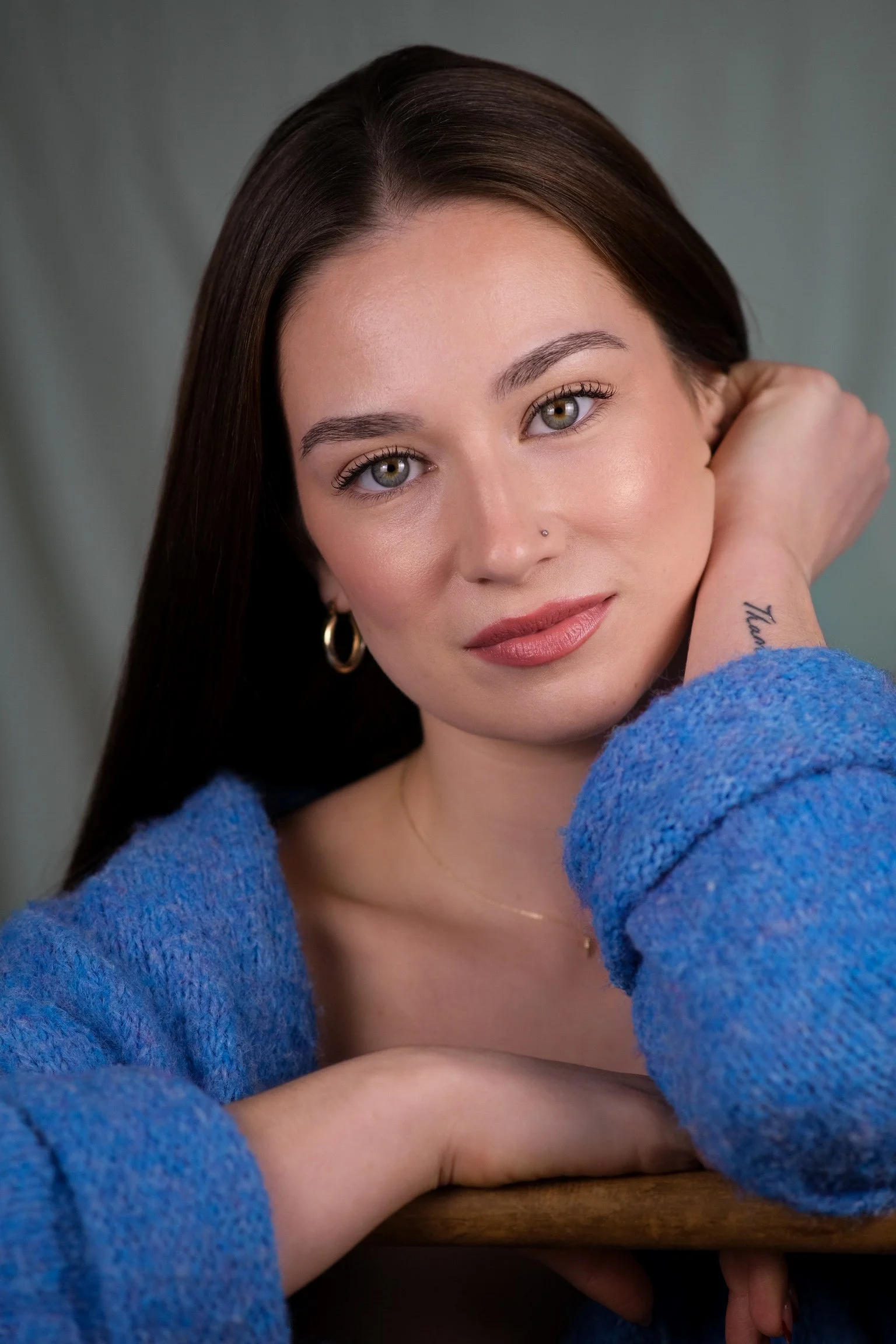 A young woman with long dark hair, green eyes, and a nose piercing, wearing a blue sweater, looking at the camera with her hand resting on her neck and her head tilted slightly. She has gold hoop earrings and a delicate necklace.