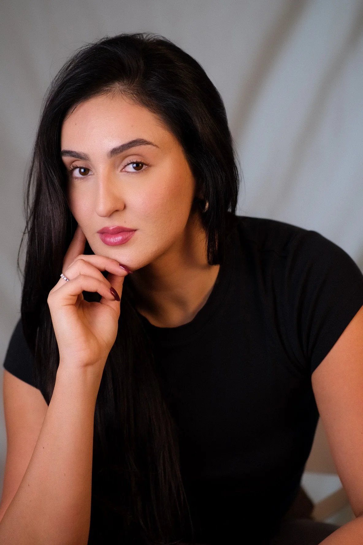 Close-up portrait of a woman with long dark hair, wearing a black t-shirt, looking at the camera with her hand resting on her chin.