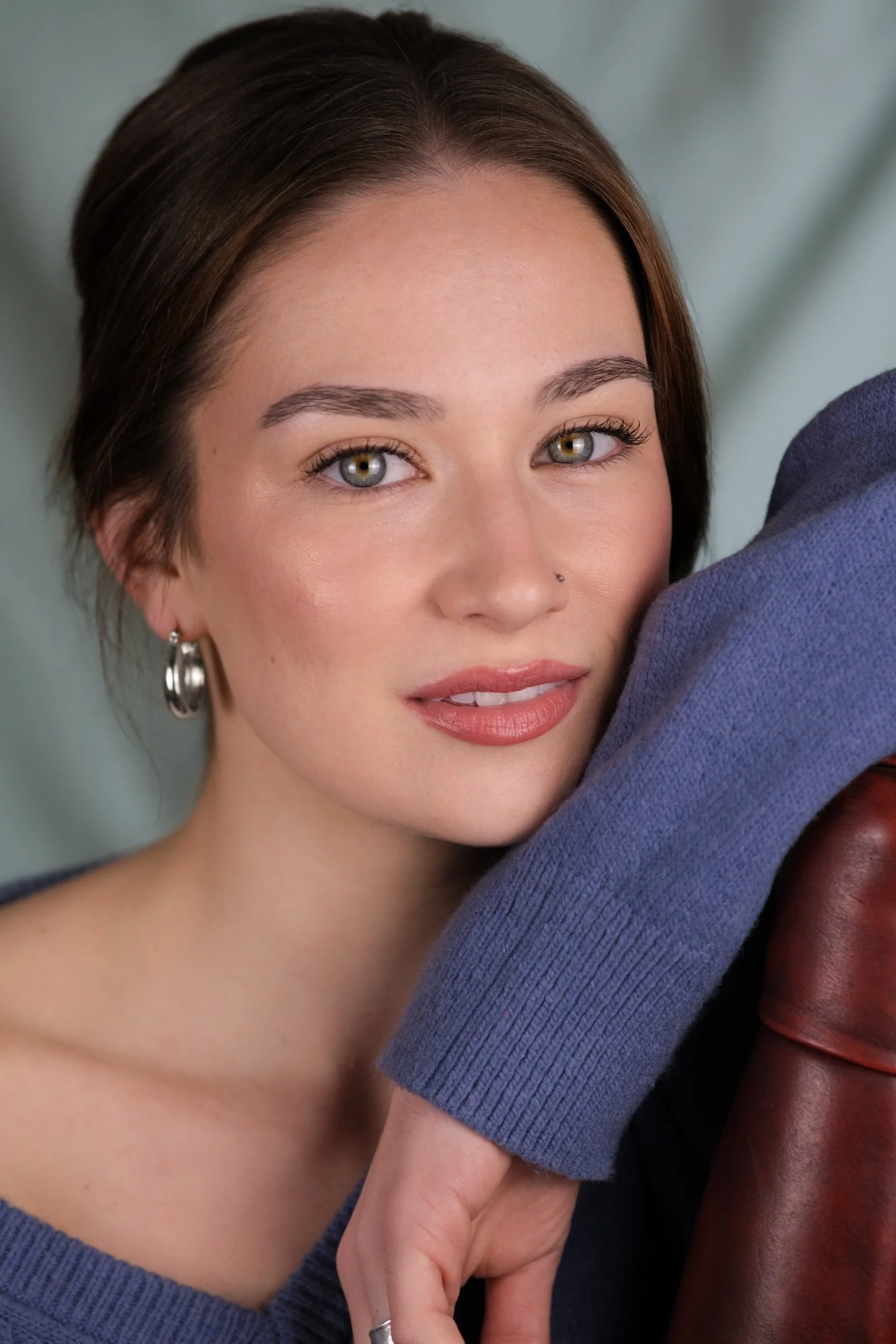 Close-up portrait of a young woman with short brown hair, gray eyes, and light makeup, wearing a blue sweater and hoop earrings, resting her face on her arm, with a soft focused background.