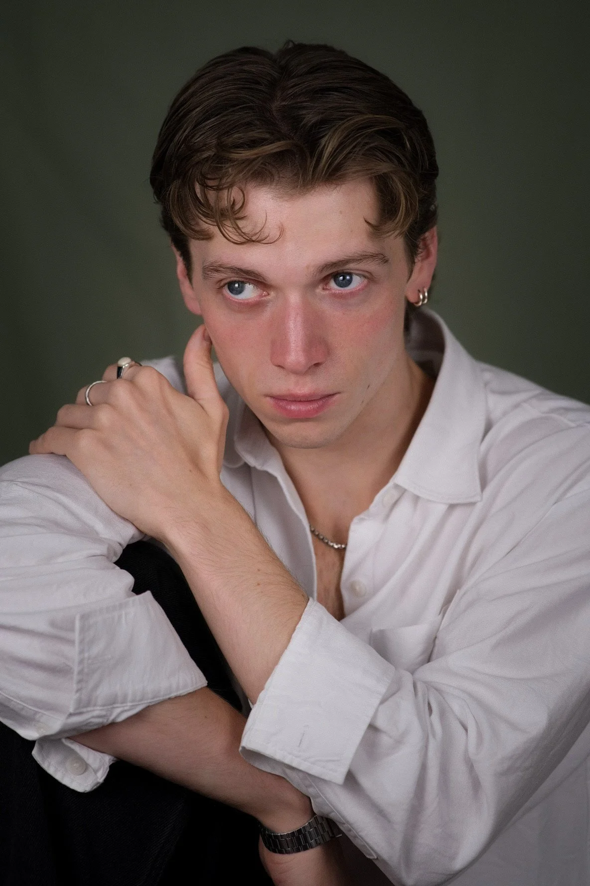 A young man with blue eyes and wavy brown hair, wearing a white shirt, earrings, and a necklace, sits with his head resting on his arm, looking thoughtfully to the side.