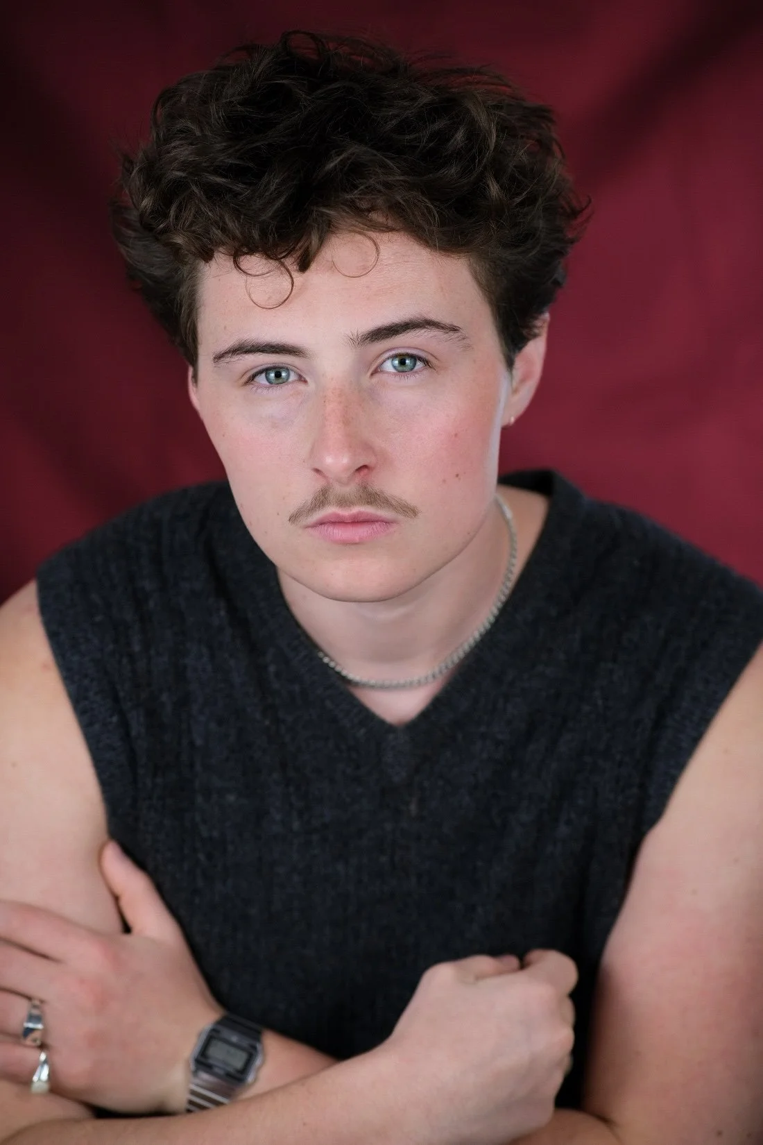 A young person with blue eyes, curly brown hair, and light skin, wearing a dark sleeveless top, a silver necklace, a watch, and rings, looking directly at the camera with arms crossed, against a reddish-burgundy blurred background.