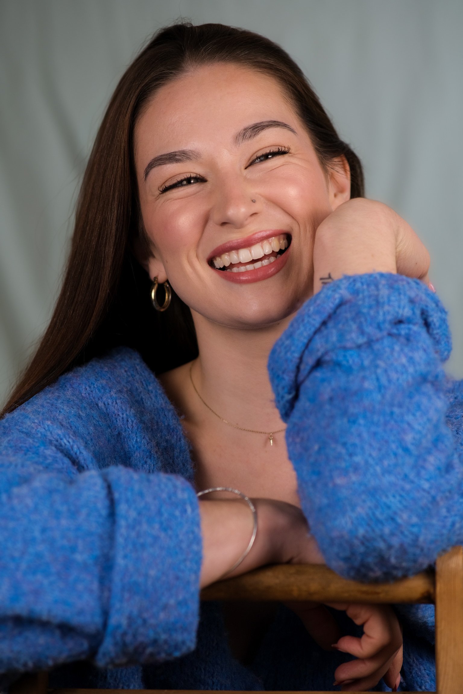 A young woman with long brown hair, smiling and showing her teeth, wearing gold hoop earrings, a thin gold necklace, and a blue fuzzy sweater, resting her chin on her arm.