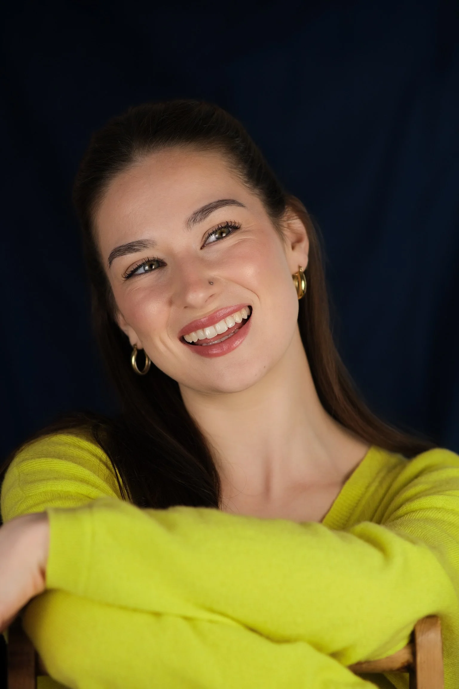 A young woman with dark brown hair smiling, wearing gold hoop earrings and a bright yellow sweater, posing with her arm resting on a wooden chair against a dark background.