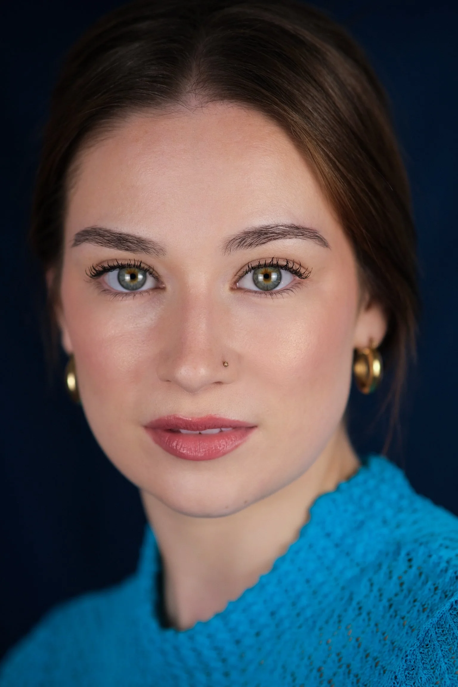 Close-up portrait of a young woman with blue eyes, short brown hair, gold hoop earrings, and wearing a blue knitted top, looking directly at the camera.