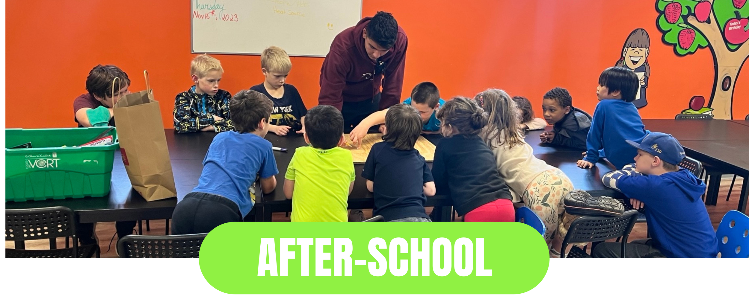 A group of young children gathered around a table with their teacher in an after-school classroom. The children are paying attention to the activity on the table, which the teacher is demonstrating. The background has bright orange walls with colorful illustrations, including a smiling girl and a tree with apples. The image has a green label at the bottom with white text that says 'AFTER-SCHOOL.'