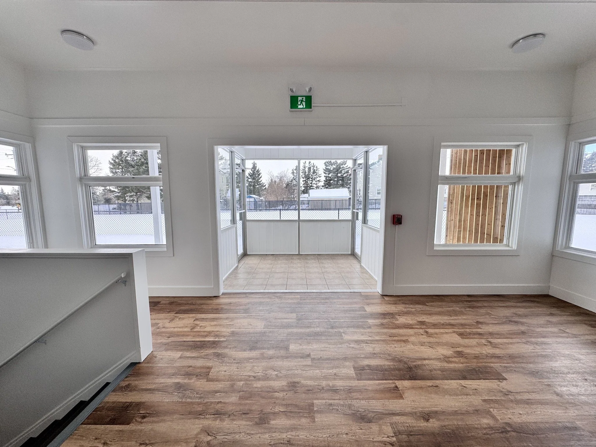 Empty room with wood flooring, white walls, large windows, and an open door leading to a screened porch with a tiled floor. Outside is snowy with trees and fences visible.