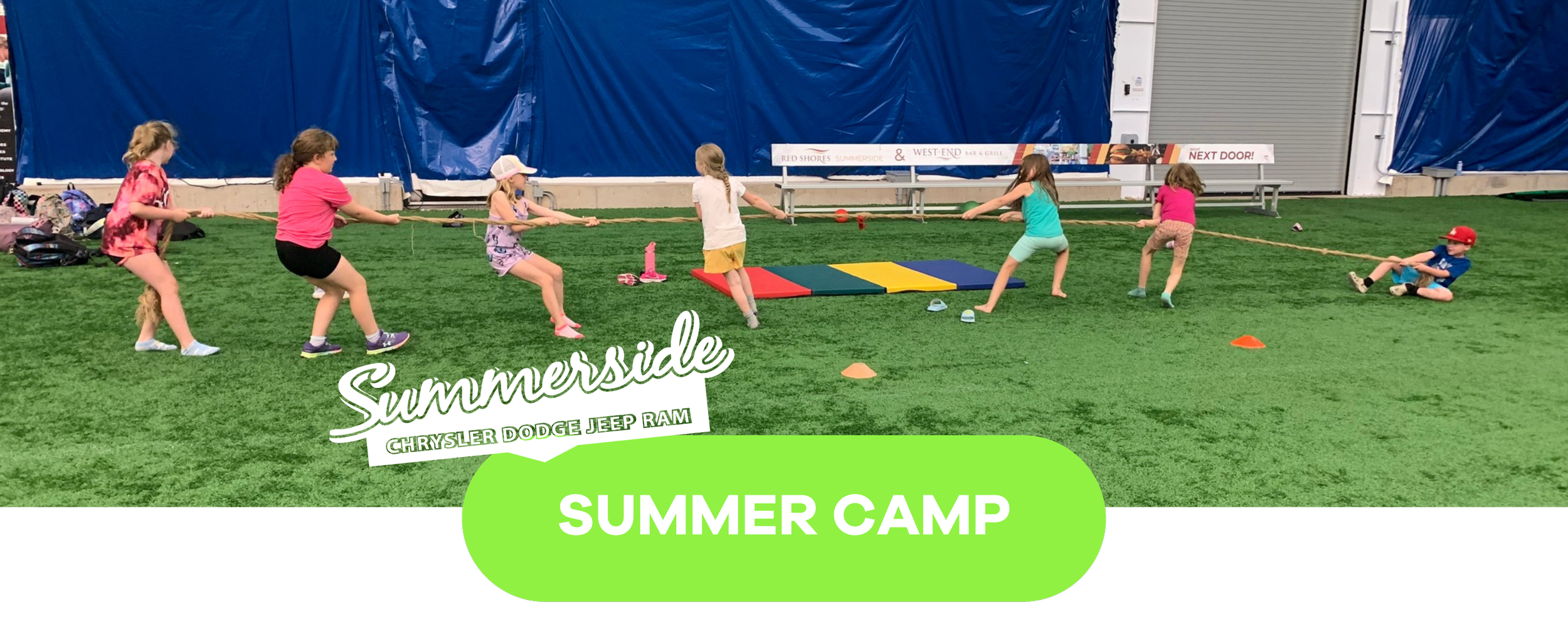 Children participating in a tug-of-war game during a summer camp indoors on a green turf field.