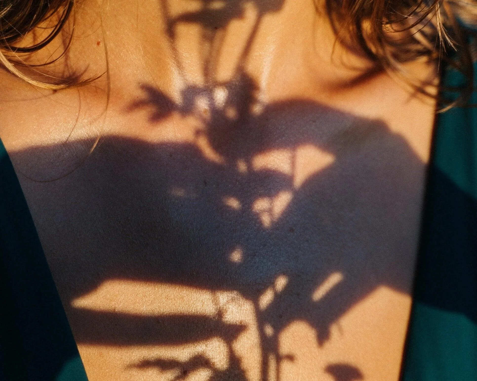 A close-up of a person's shoulder with sunlight casting a shadow of a plant's leaves and branches on their skin.