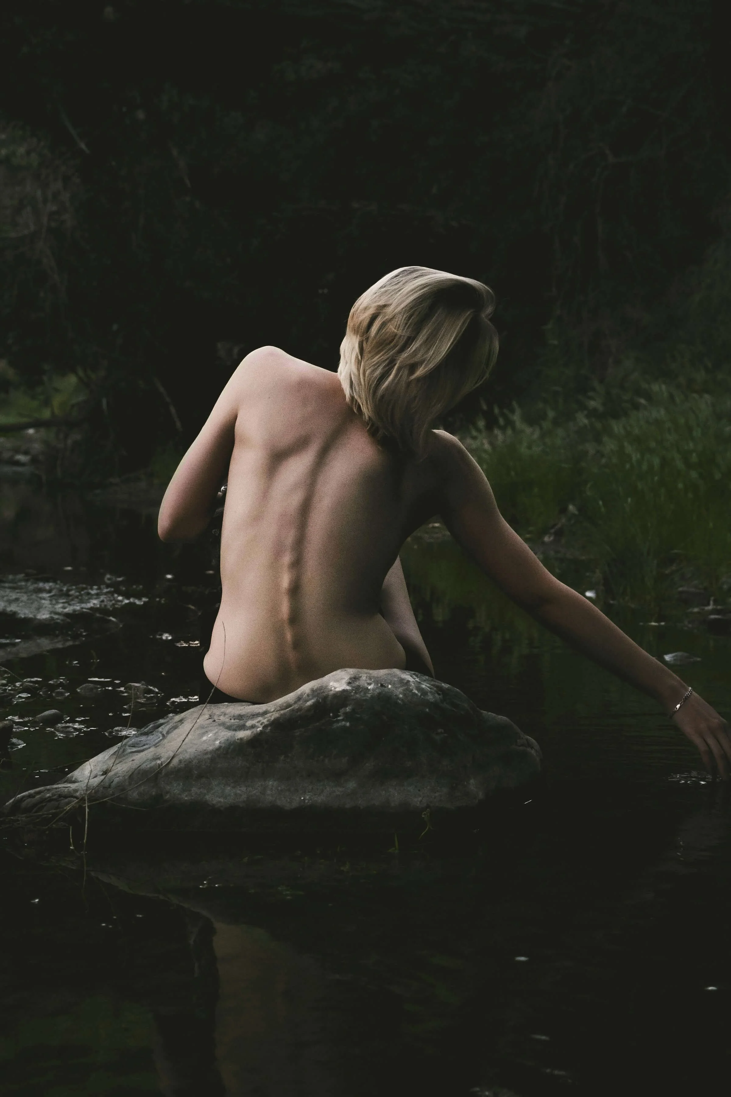 A nude woman with blonde hair sitting on a rock in a dark, forested stream, with her back to the camera and one arm extended into the water.