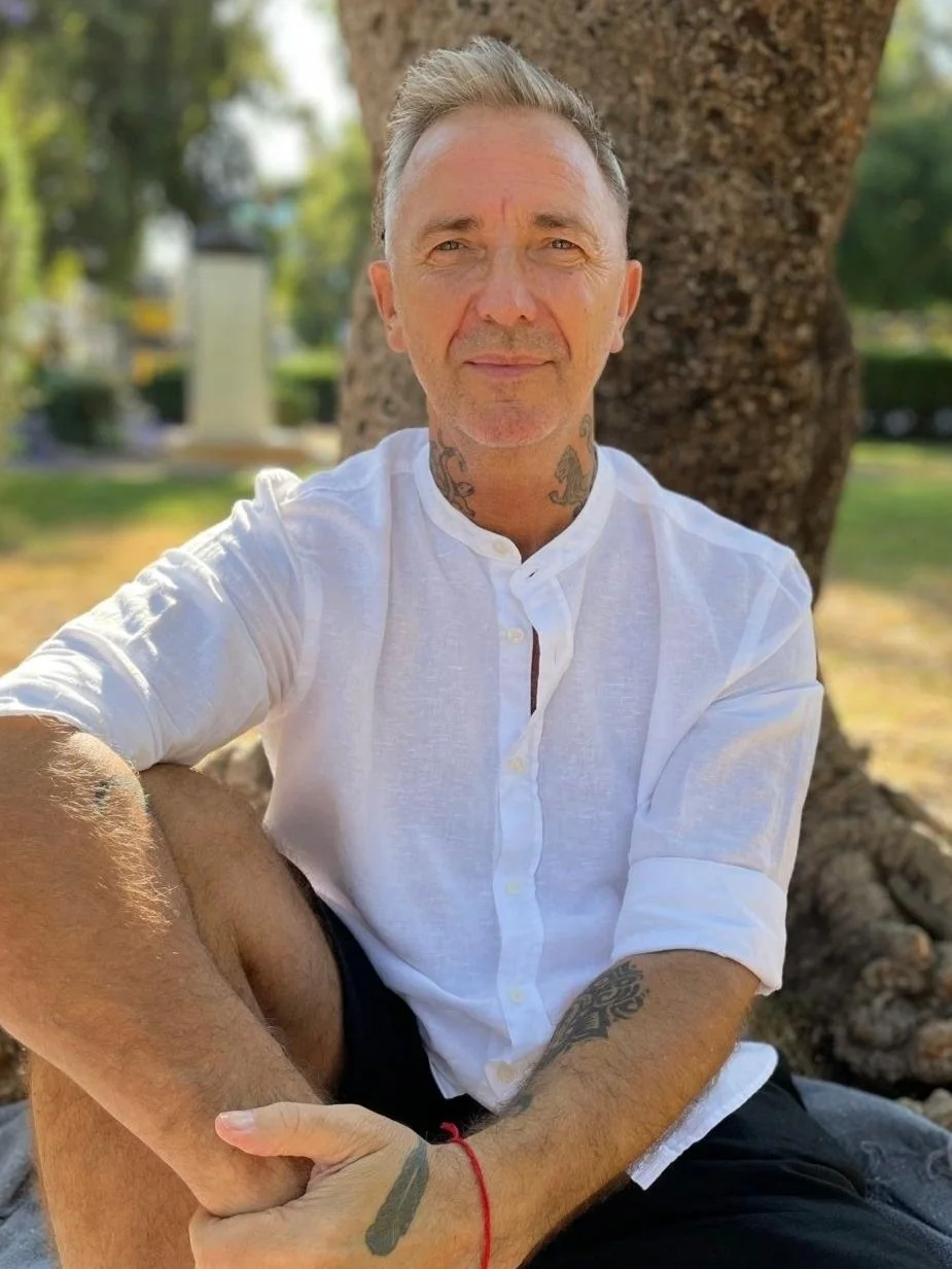 A man sitting outdoors in front of a tree, wearing a white button-up shirt with the sleeves rolled up, and black shorts. He has tattoos on his neck and arm, and gray hair. The background shows green grass, blurred trees, and some distant structures.