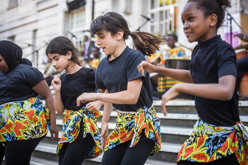 A group of girls dances wearing black outfits with colourful floral skirts on top