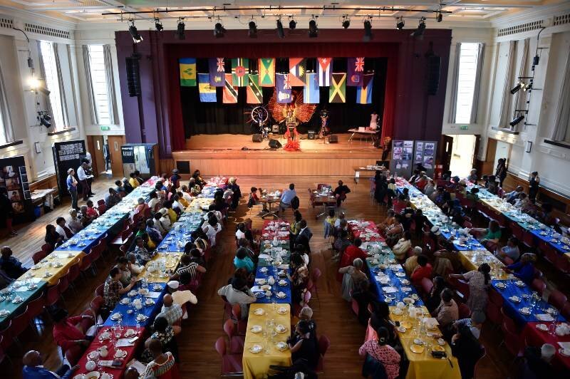 People seated at a series of long tables with a stage in the background of the image. On the stage are displayed several flags hanging down.