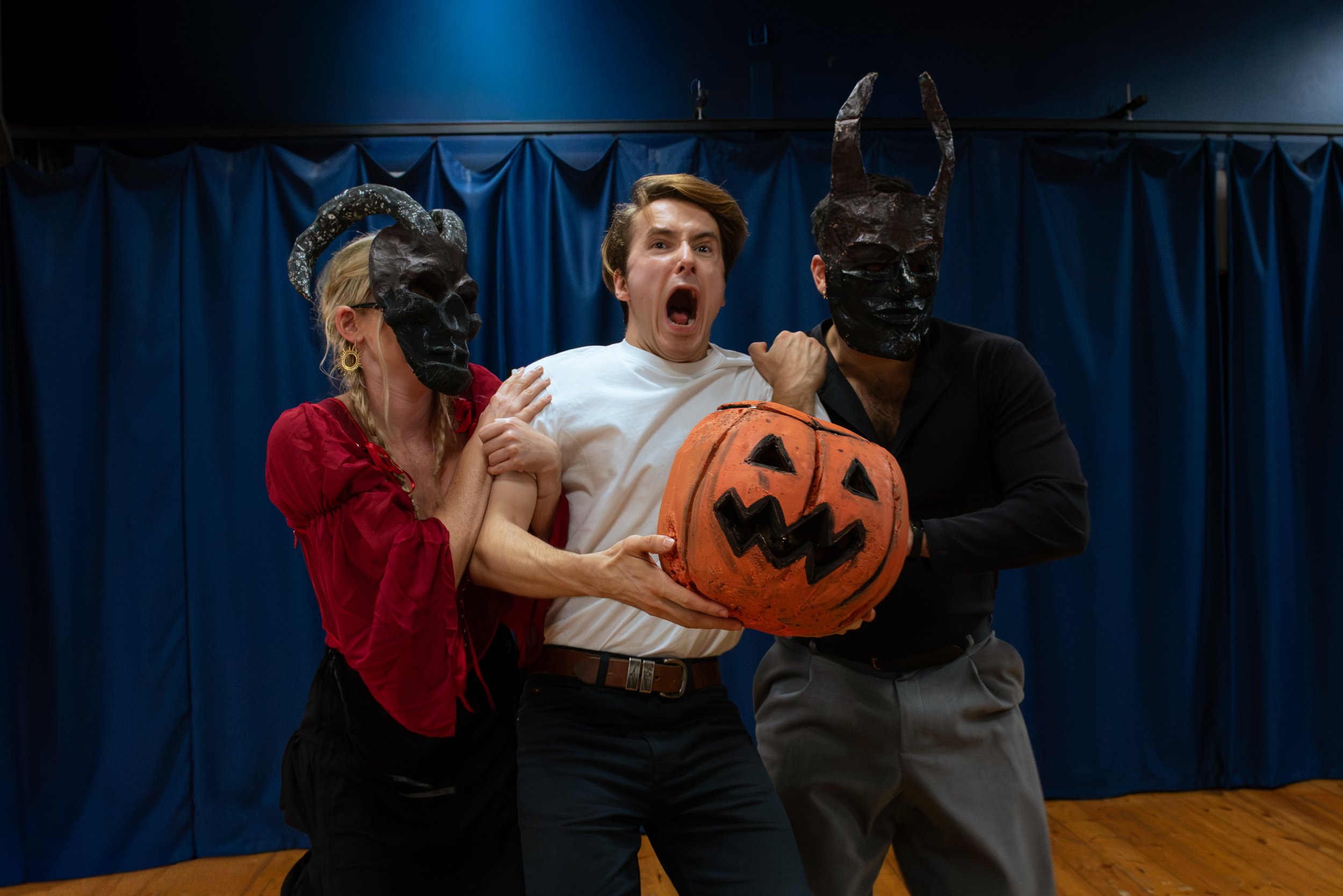 Two performers wearing masks frighten a third performer who holds a pumpkin.