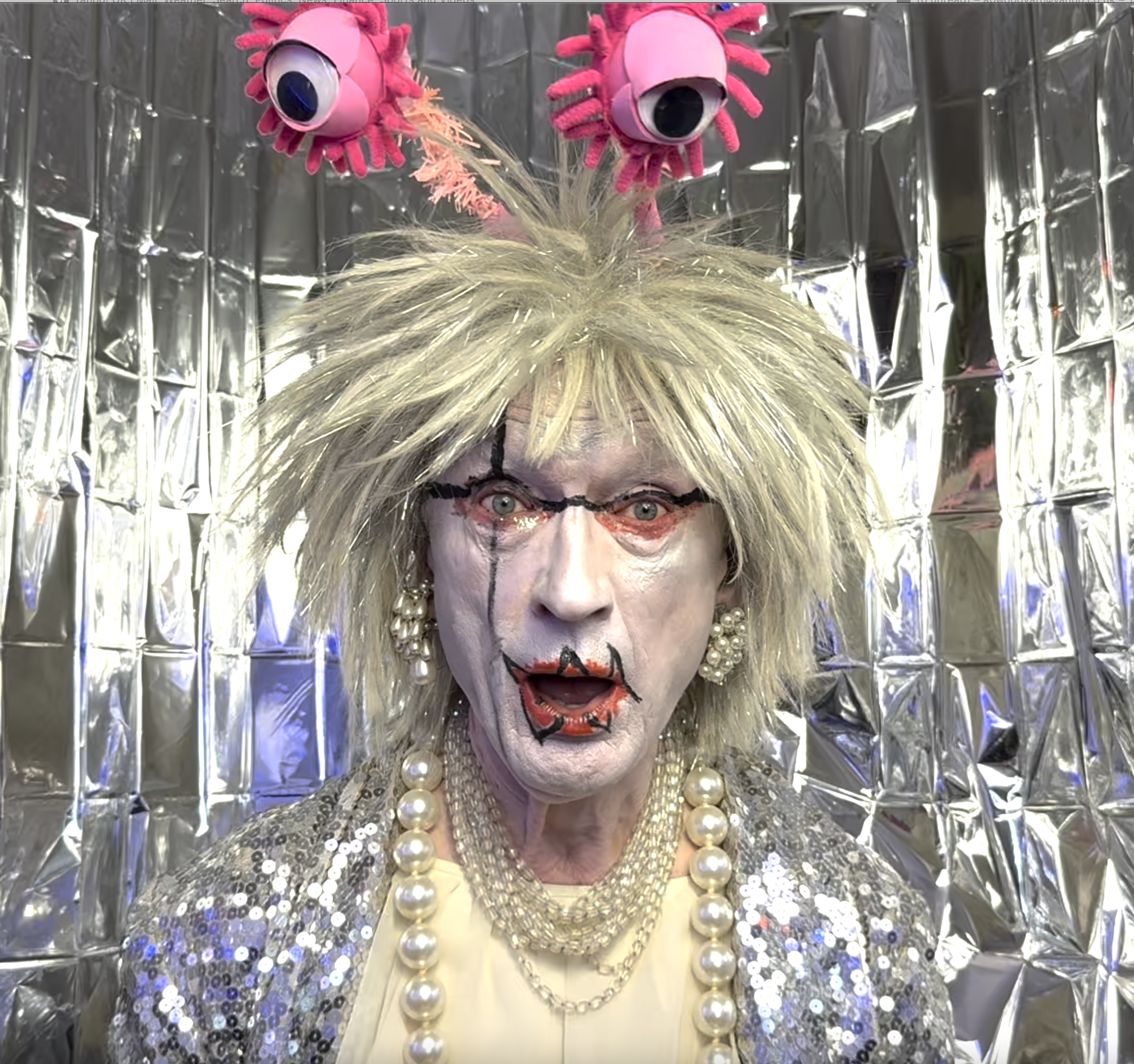 A drag queens face infant of a silver background, they have a full face of make up, futuristic hair and goggly eye headpiece