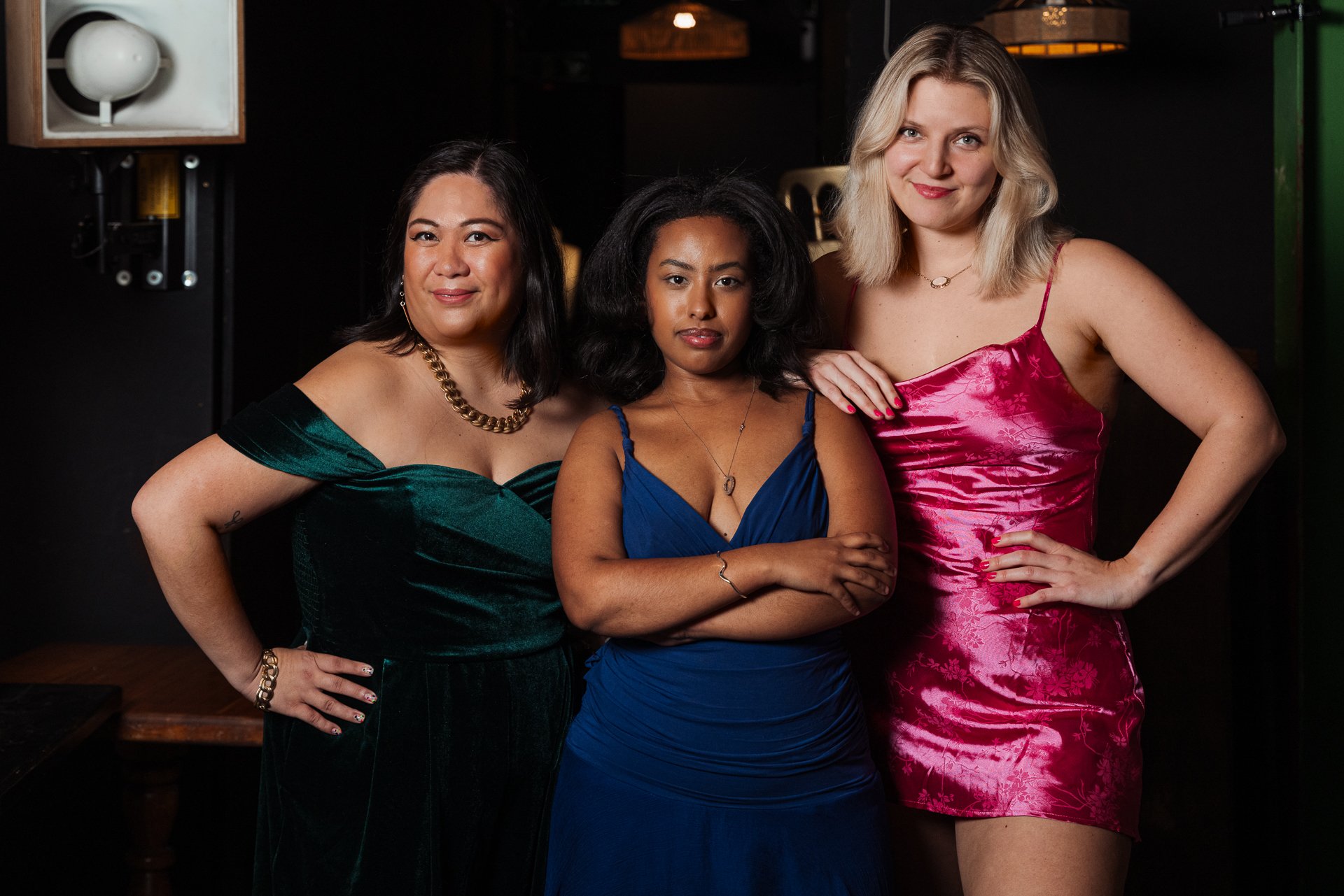 three women of different ethnicities stand in jewel coloured dresses looking at the camera, they have mischievous smirks