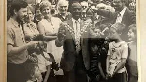 A black and white photograph of a man in a suit wearing a mayoral necklace of medallions, he is surrounded by a crowd of people