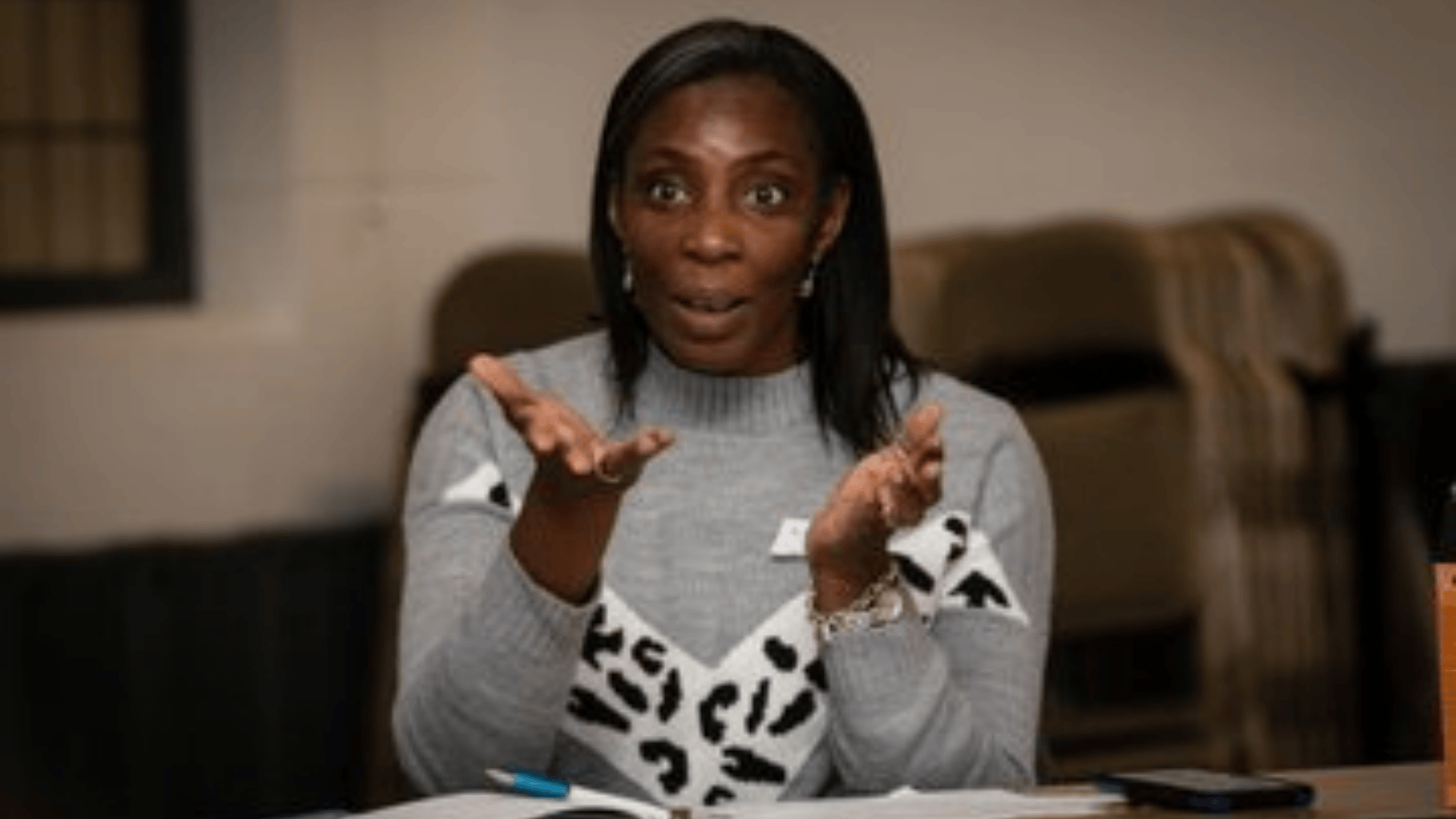 A Black woman with dark straight hair wears a grey jumper and gesticulates, as if instructing a group of people