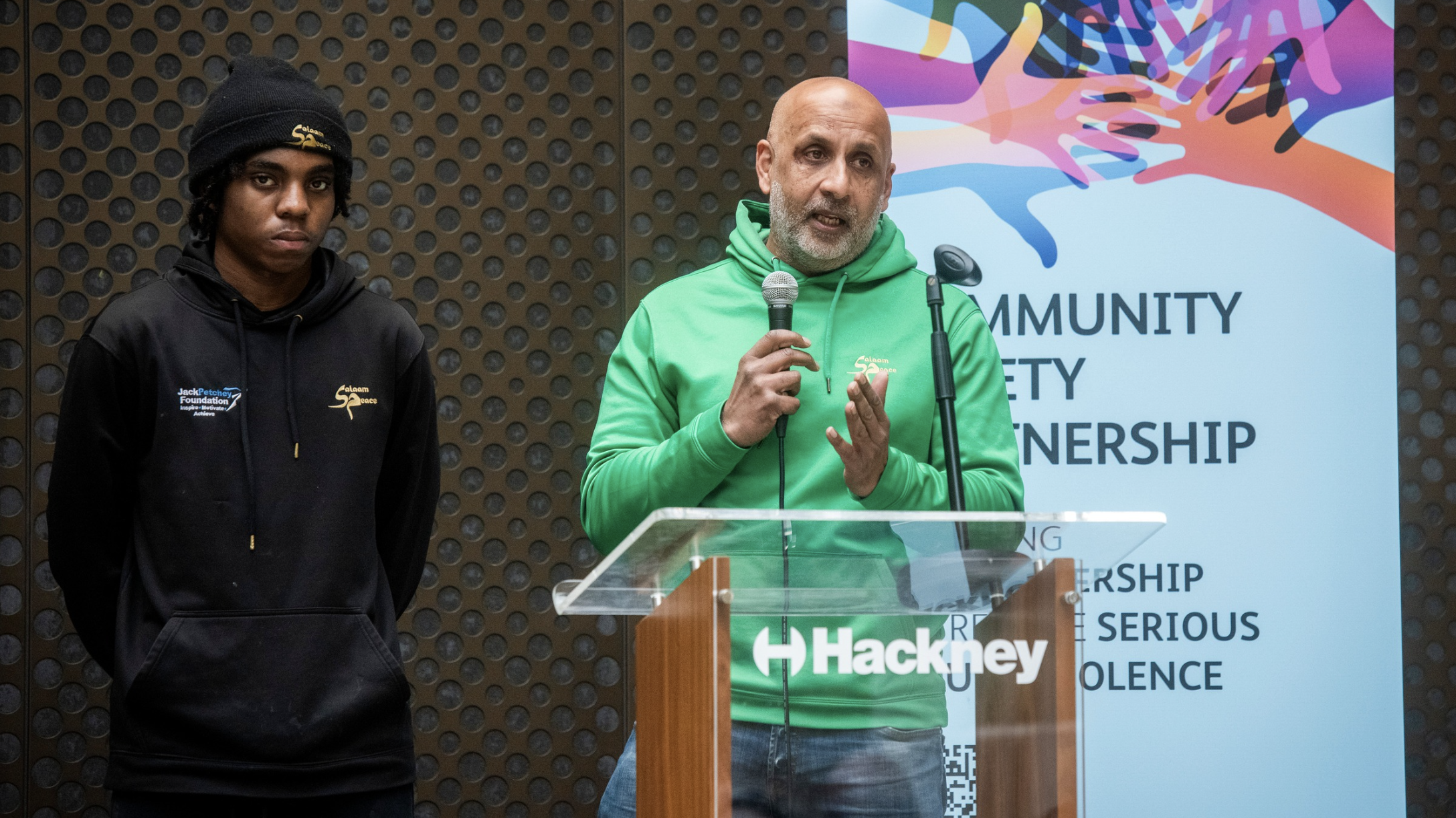 Sab Bham wears a green hoodie and holds a microphone. A young person stands next to him