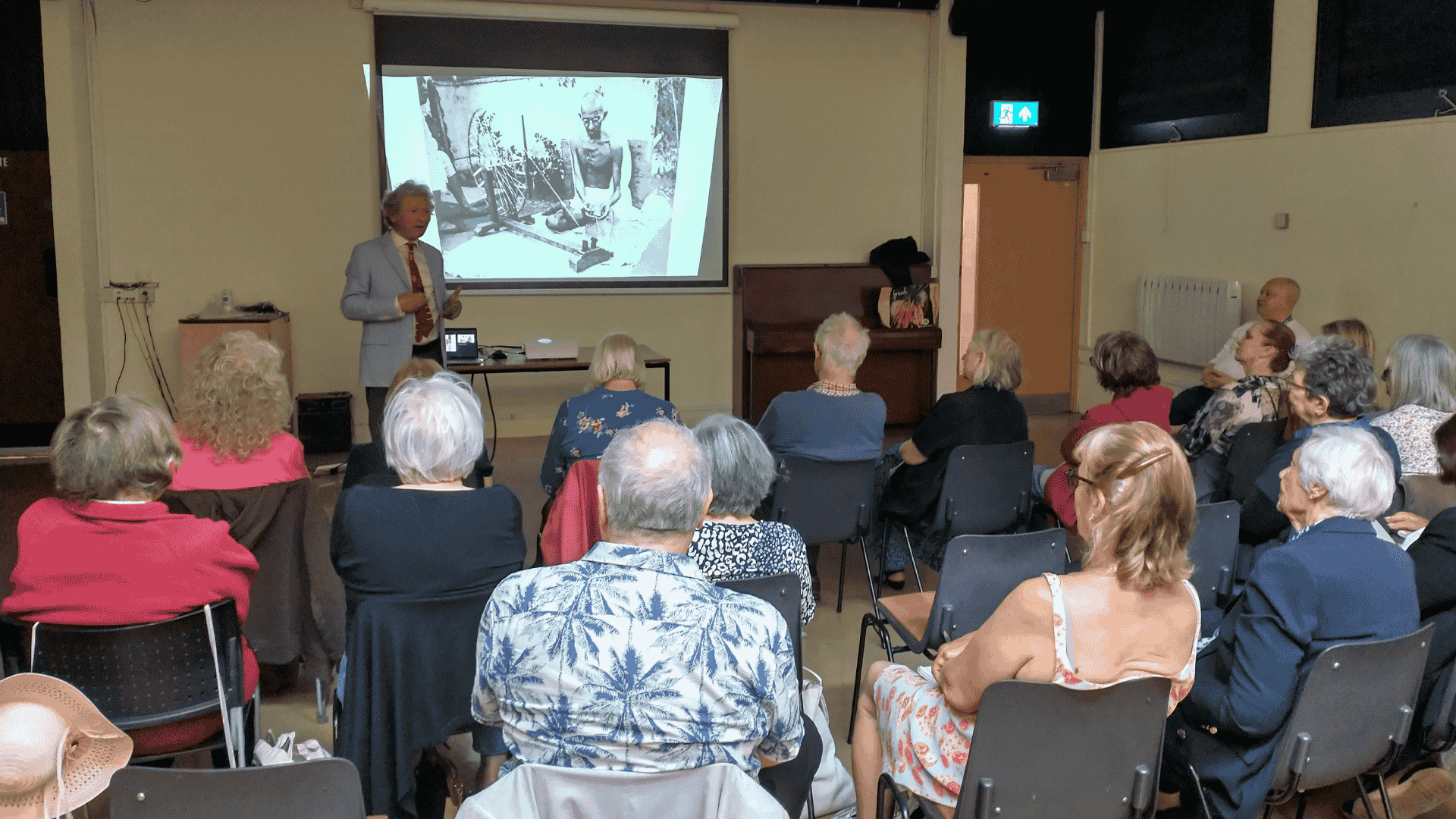 A group of people seated while a man stands at the front of the room in front of a projected image on a presentation