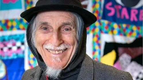 An older man with a smile on his face stands in front of a blanket tapestry, he wears a hood pulled with a black pork pie style hat over it
