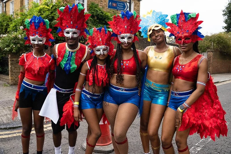A group of young people pose in a line in the street. They are wearing carnival style costumes with red feather headresses and masks, blue shiny shorts and red crop tops.