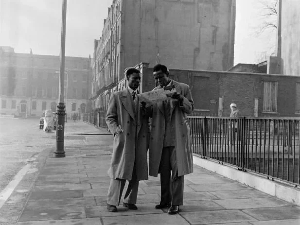 A black and white photo of two men on a London street wearing long coats and looking at a newspaper